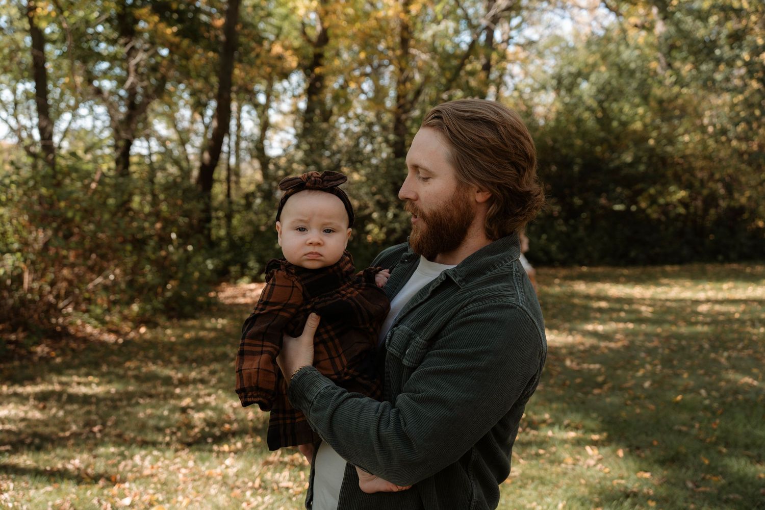A bearded father holds his infant baby dressed in a brown bear costume outdoors in a sunlit autumn park.