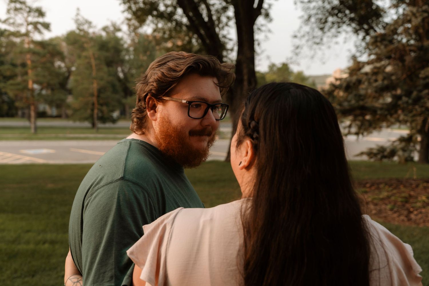 Couple sharing a romantic moment in a park at golden hour, man with glasses and red beard facing woman with dark hair.