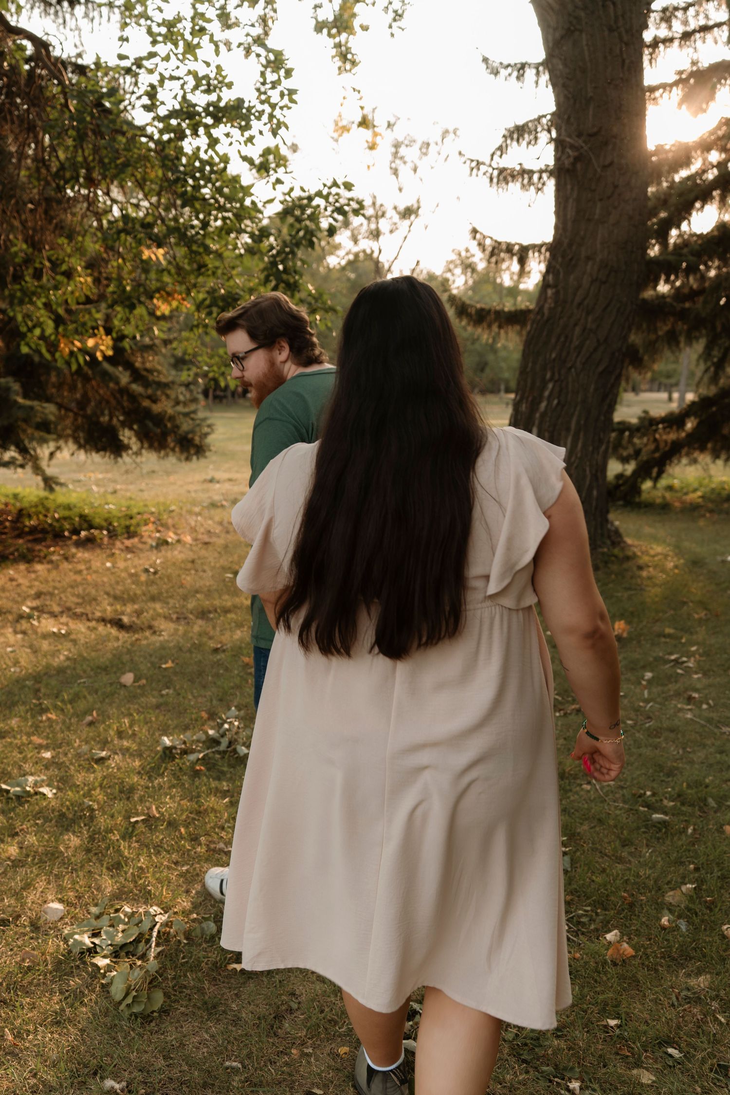Couple walking hand in hand through a sunlit park, woman in cream dress with long dark hair, man in green shirt.
