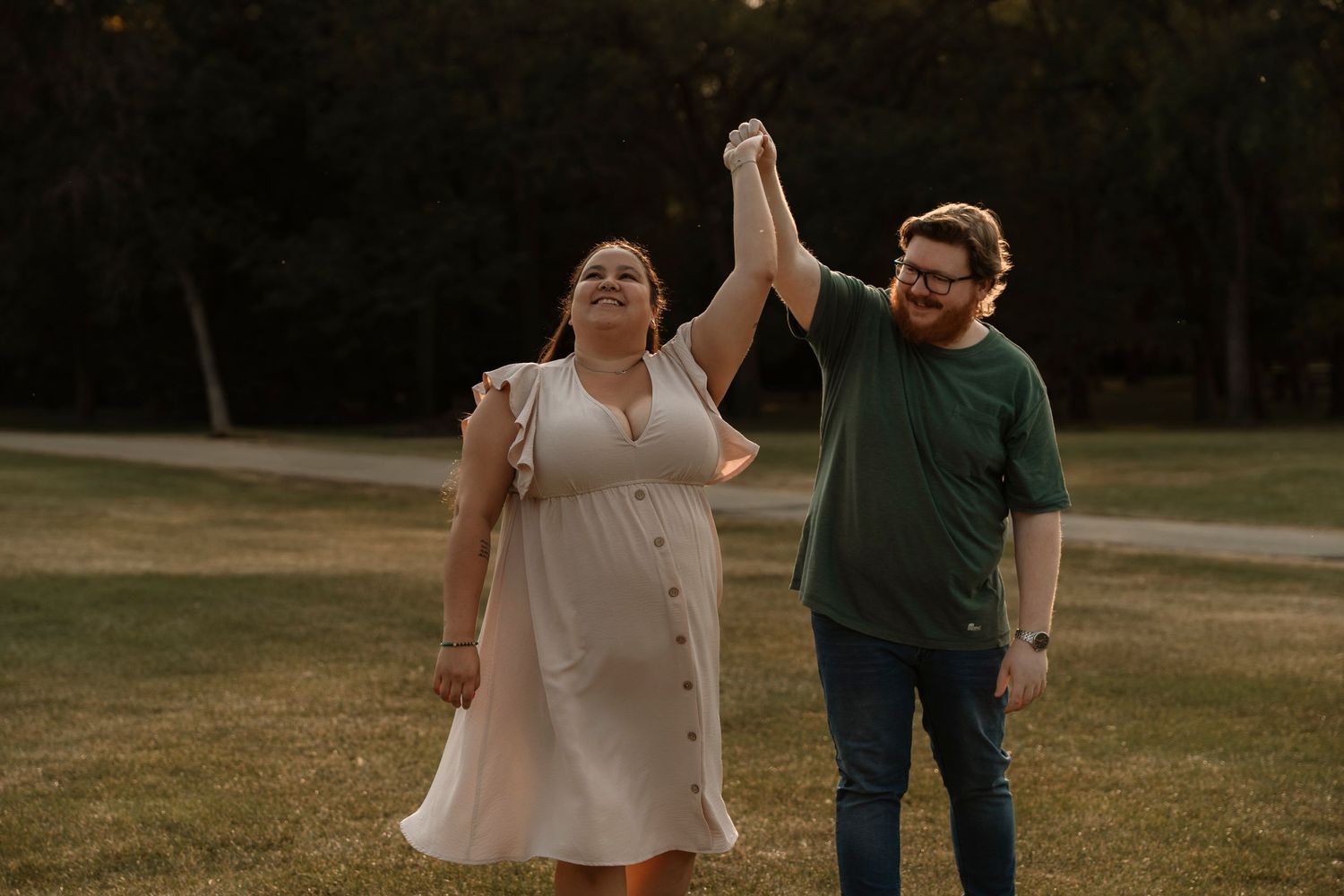 Couple dancing outdoors at sunset, woman in white dress and man in green shirt holding hands raised in a park.