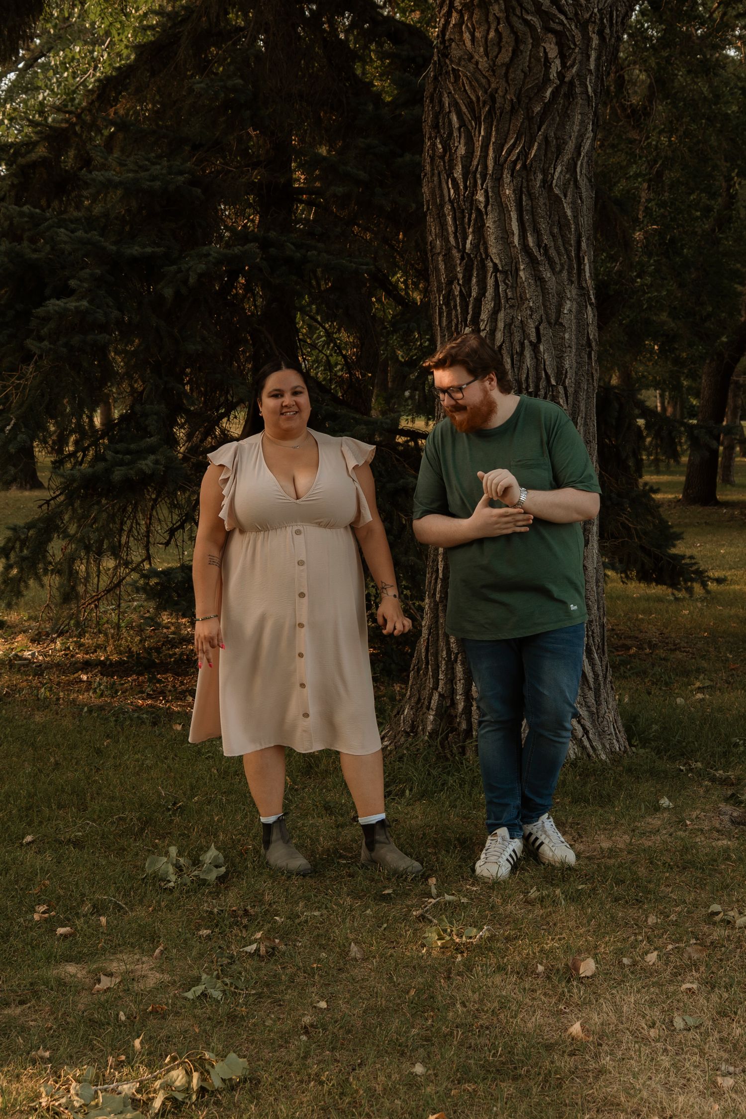 A couple stands together near a large tree in a park, woman in a beige dress, man in a green shirt and jeans.