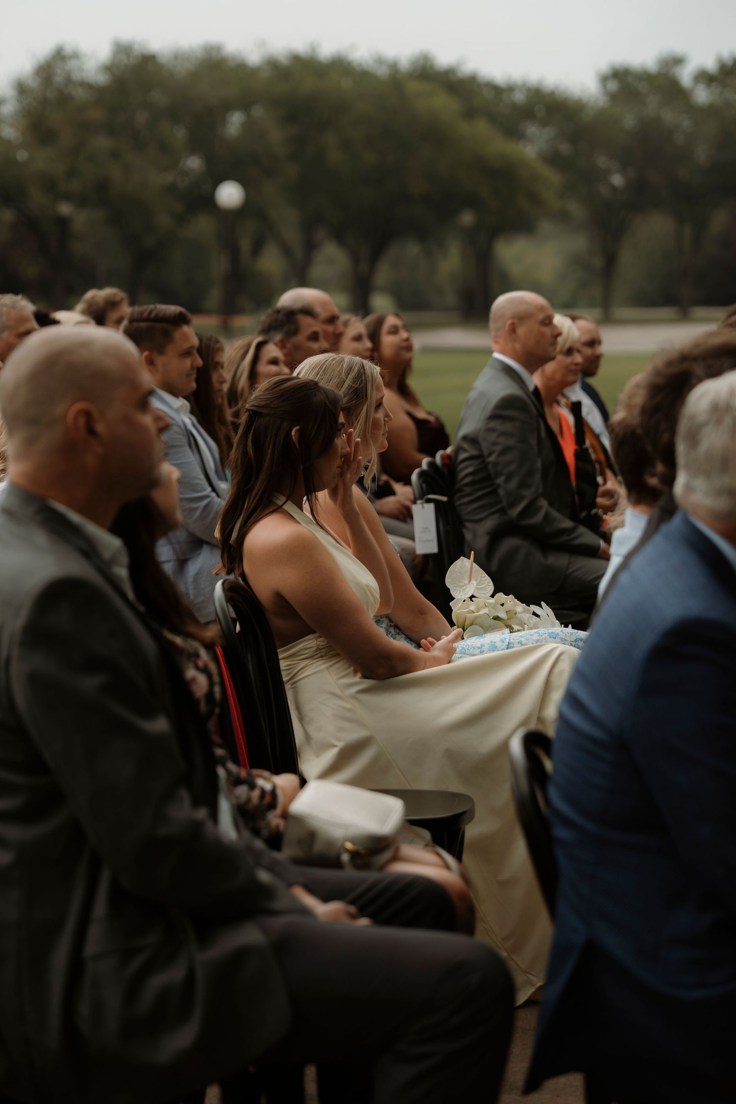 Wedding guests seated outdoors during a ceremony, dressed in formal attire, surrounded by lush green trees.