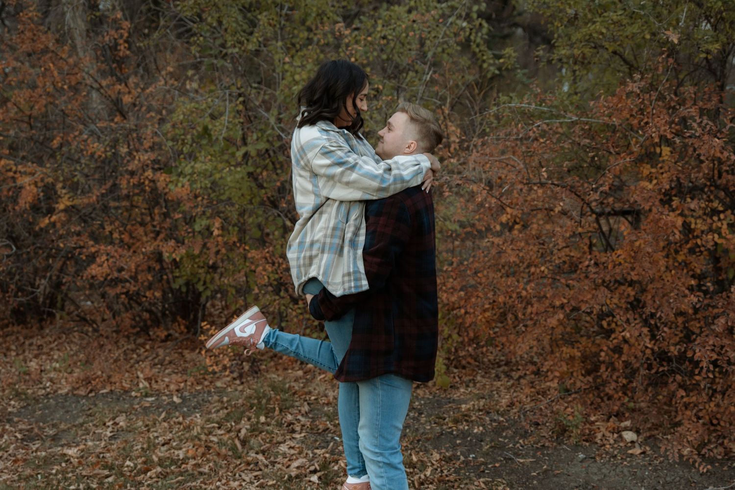 Couple embracing in autumn forest, man lifting woman among orange and red foliage in a romantic outdoor setting.