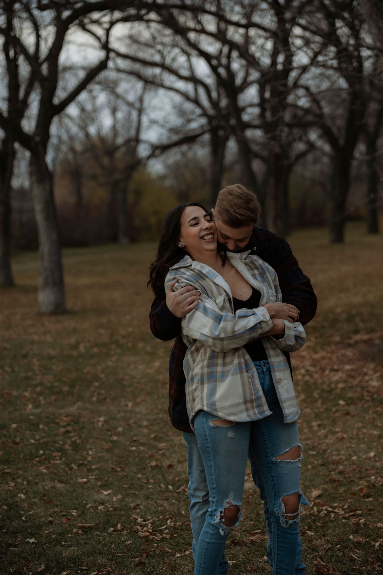 A couple embraces and laughs together in an autumn park, surrounded by bare trees.