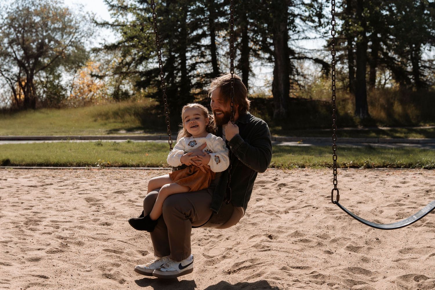 A bearded father sits on a swing holding his smiling toddler at a sandy playground surrounded by trees.