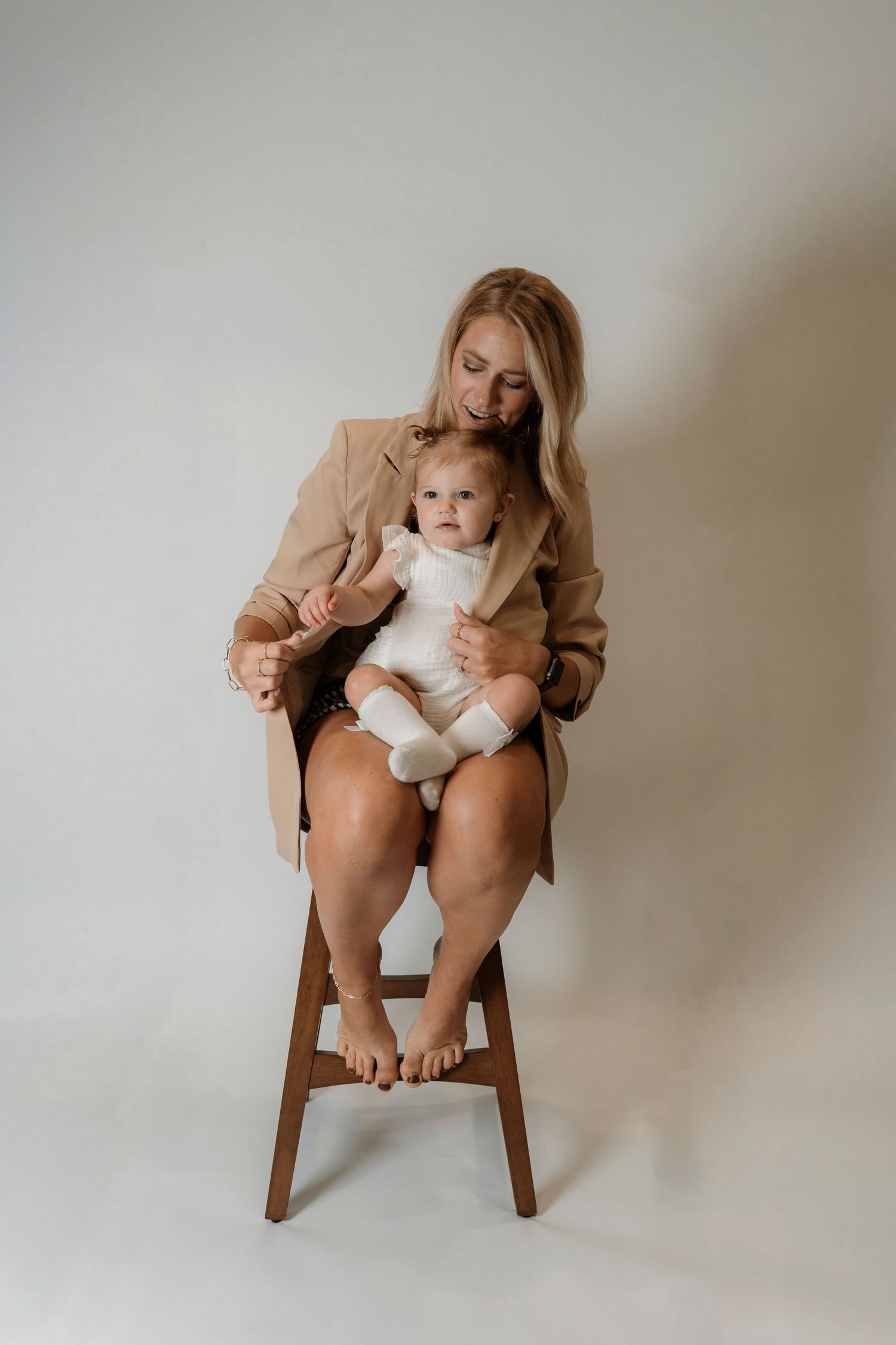 A blonde woman in a tan blazer sits on a wooden stool holding a baby in a white onesie against a light background.