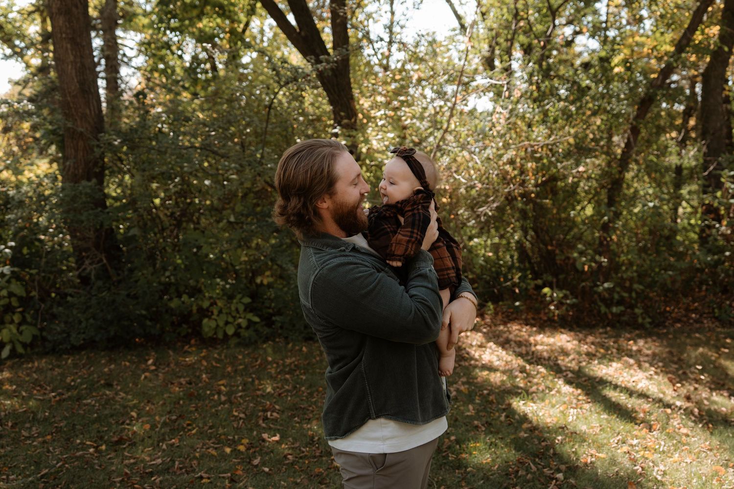 A father lifts his baby in a sunlit park surrounded by lush green trees during a family outdoor photo session.