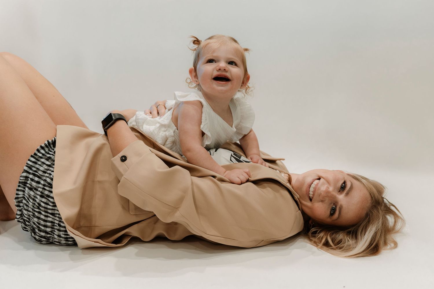 A smiling blonde woman lying on the floor wearing a tan coat with a happy baby crawling on her back.