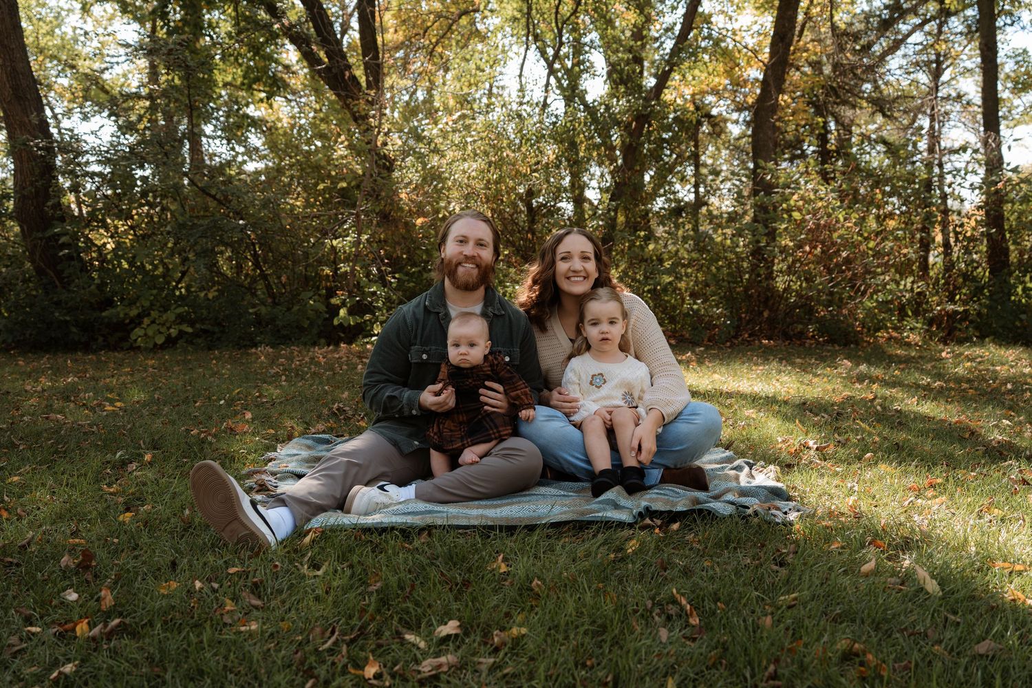 Happy family of four sitting on blanket in autumn park, smiling together.