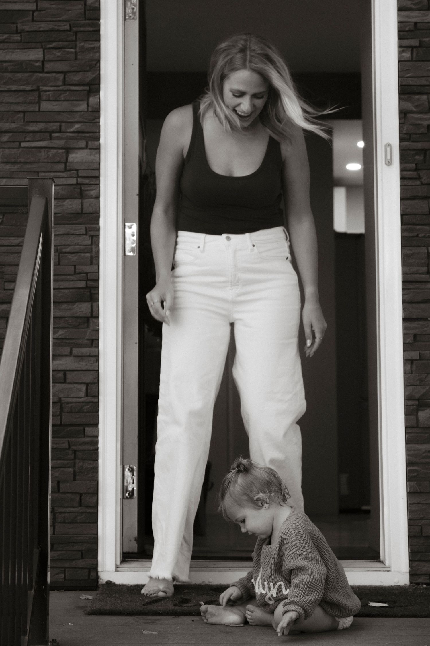 Black and white photo of a smiling blonde woman in white jeans standing in a doorway watching a toddler play on the ground.