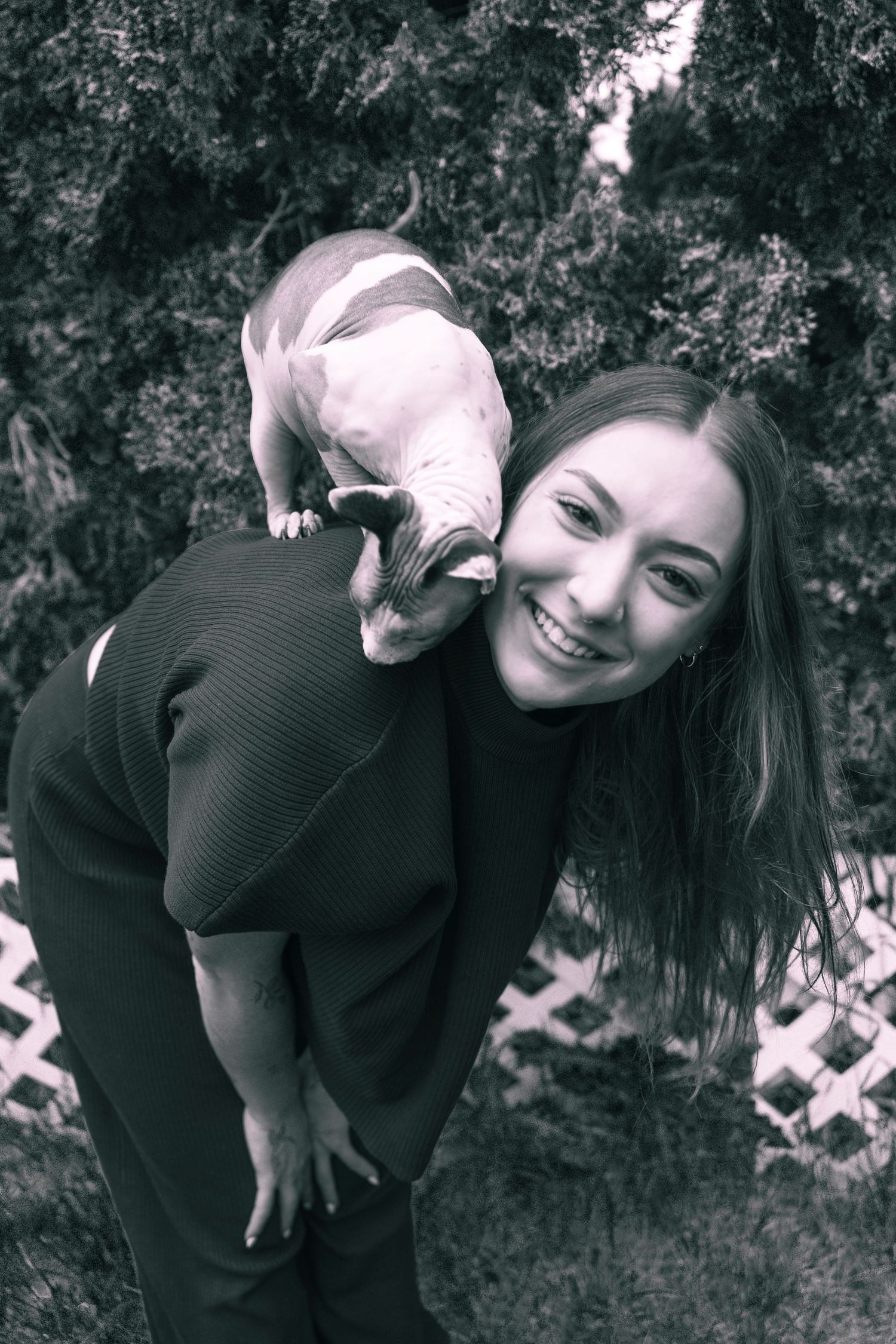 Black and white photo of a smiling young woman bending forward with a small puppy perched on her back outdoors.