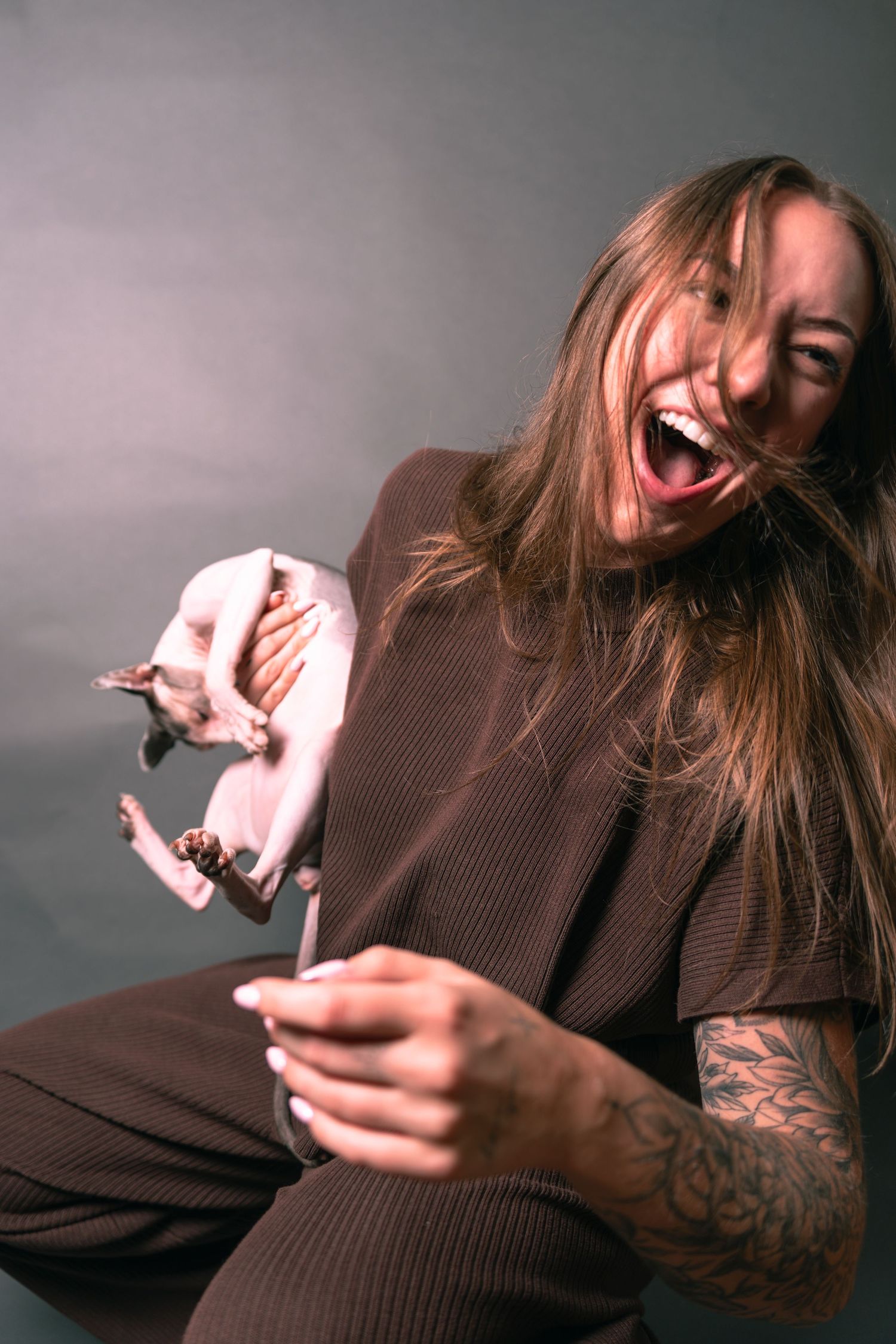 Laughing woman with tattoos holding a hairless Sphynx cat in a studio setting wearing a brown outfit.