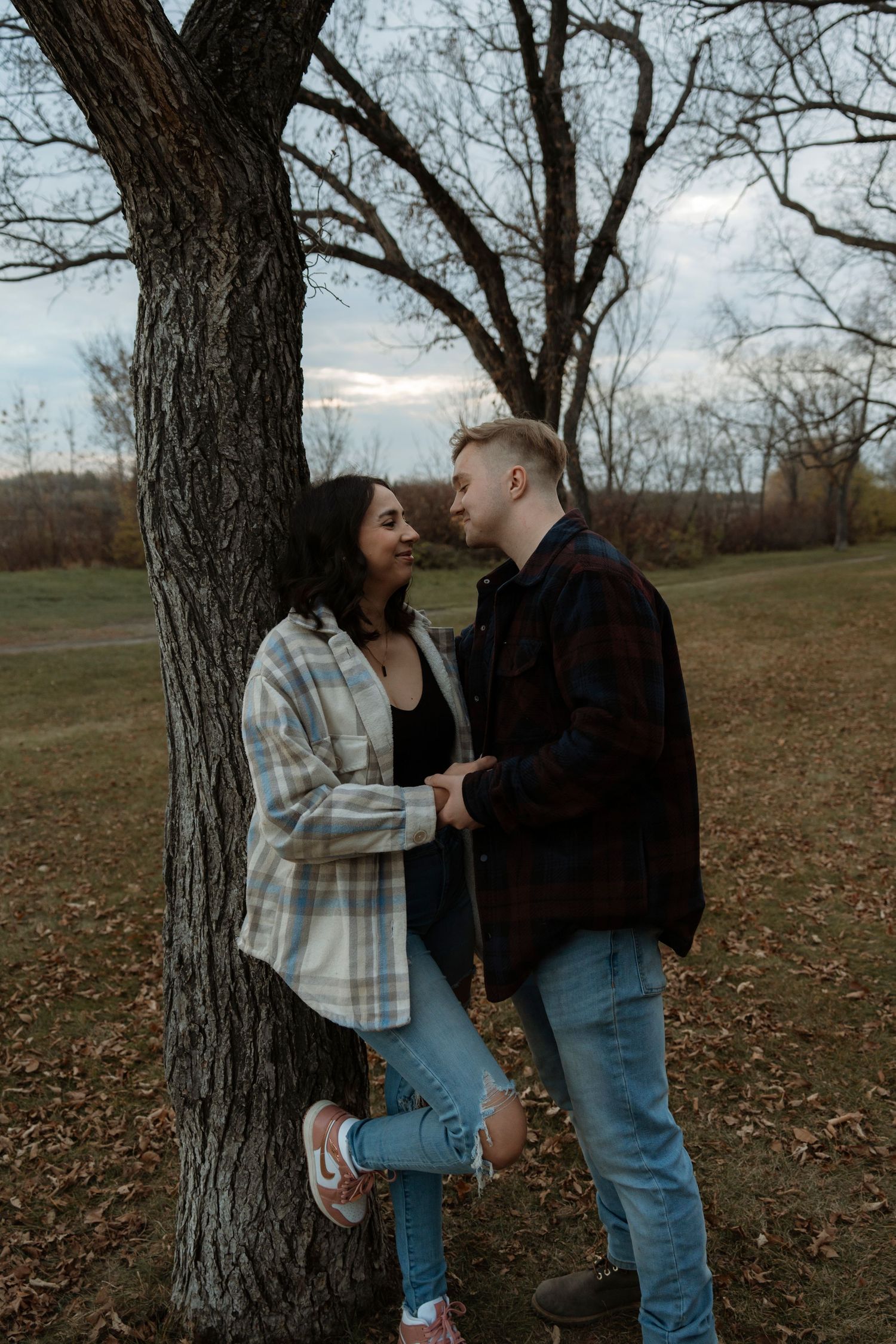 A couple shares a romantic moment leaning against a tree in an autumn park, holding hands and gazing at each other.