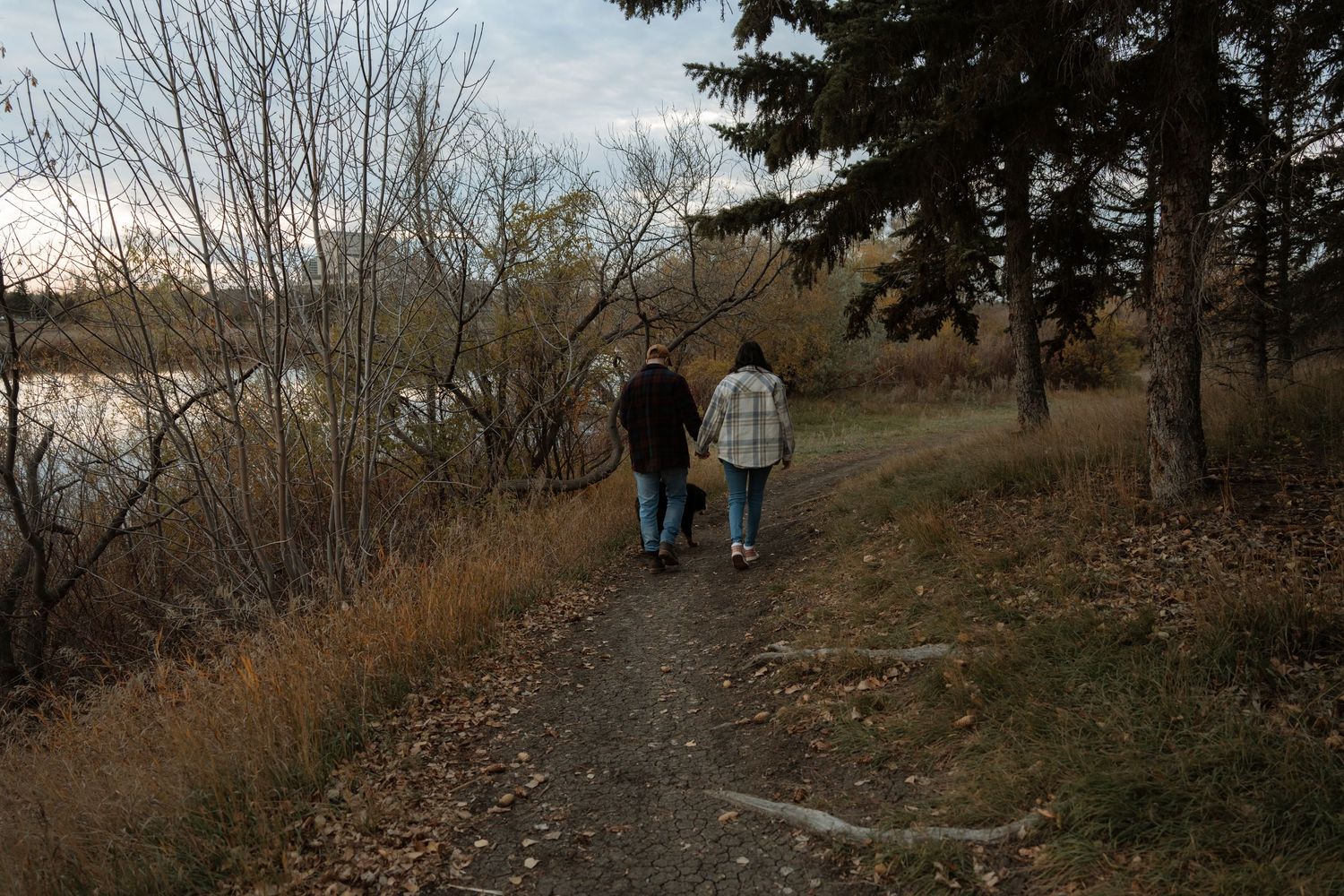 Couple walking hand in hand on a scenic autumn trail beside a calm river surrounded by bare trees.