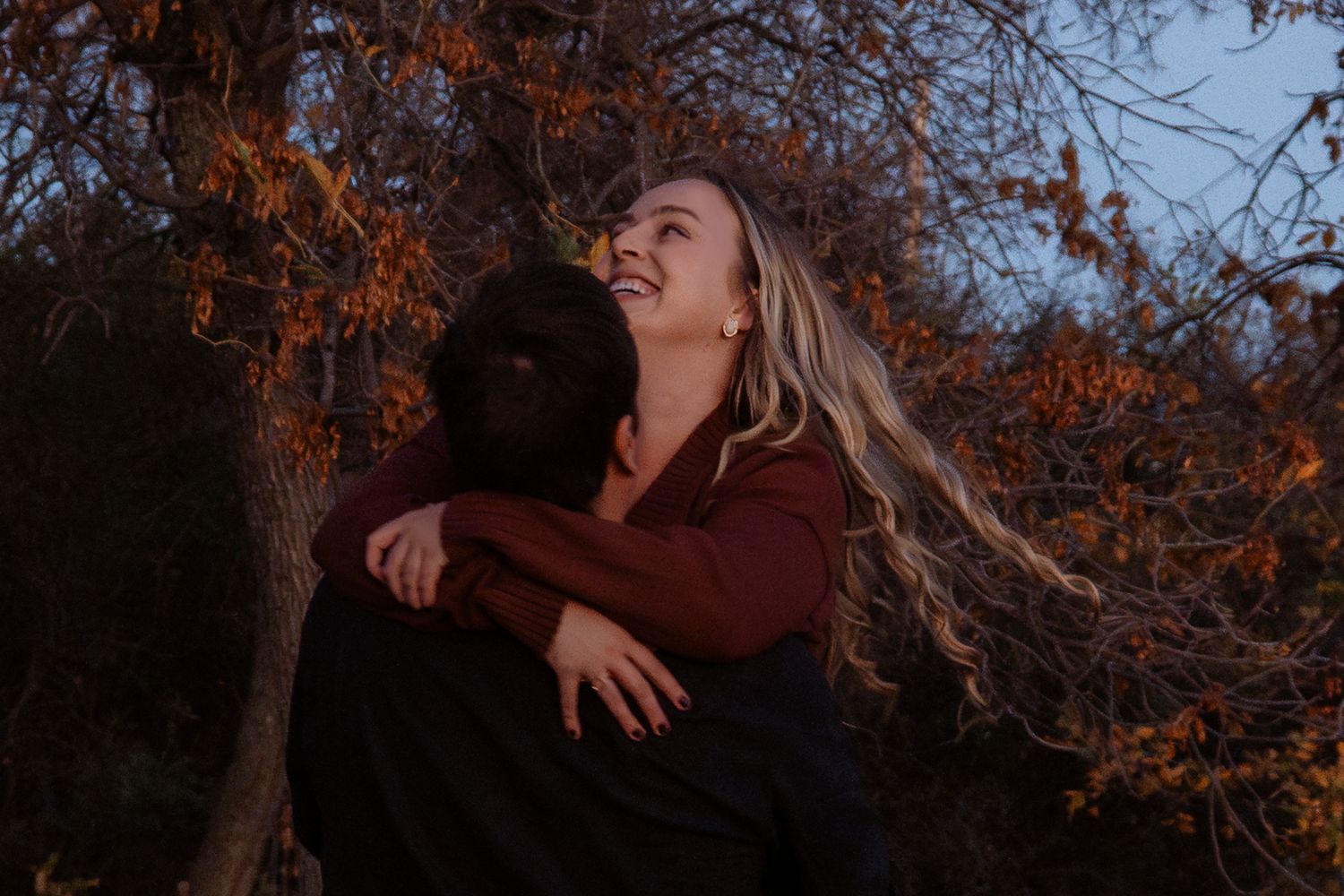 A smiling blonde woman embraces a partner near autumn trees at dusk in a romantic outdoor portrait.