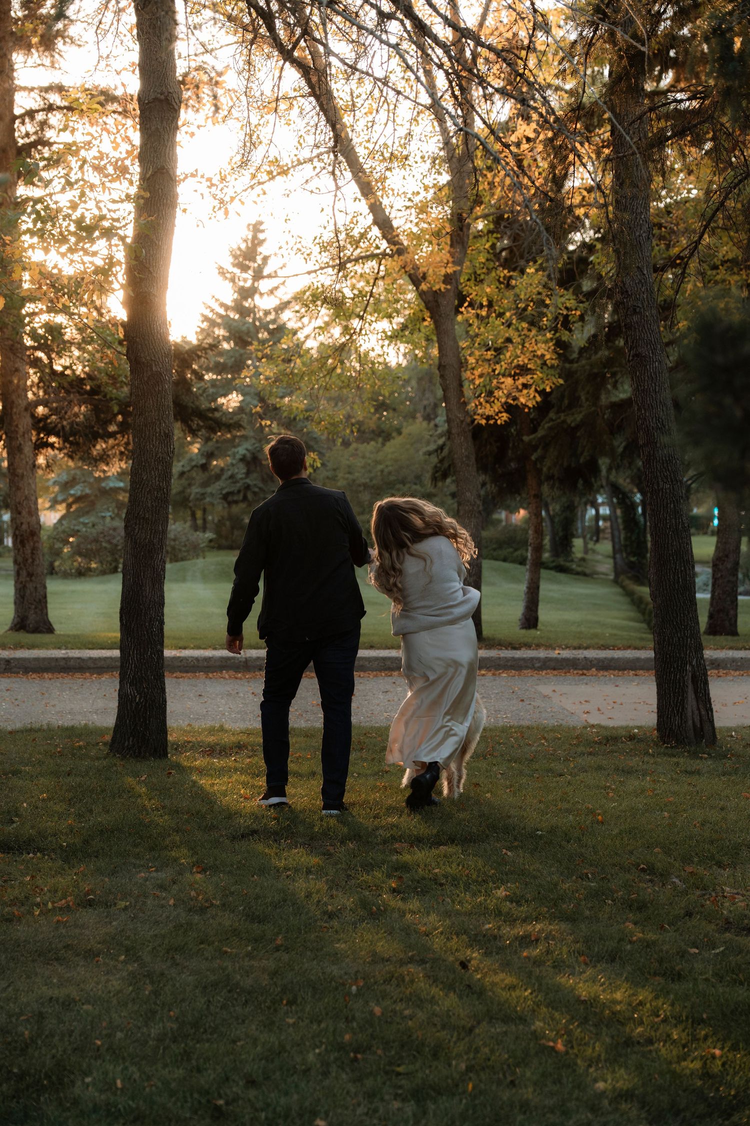 Couple walking hand in hand through a sunlit park at golden hour, woman in white dress and man in dark suit.