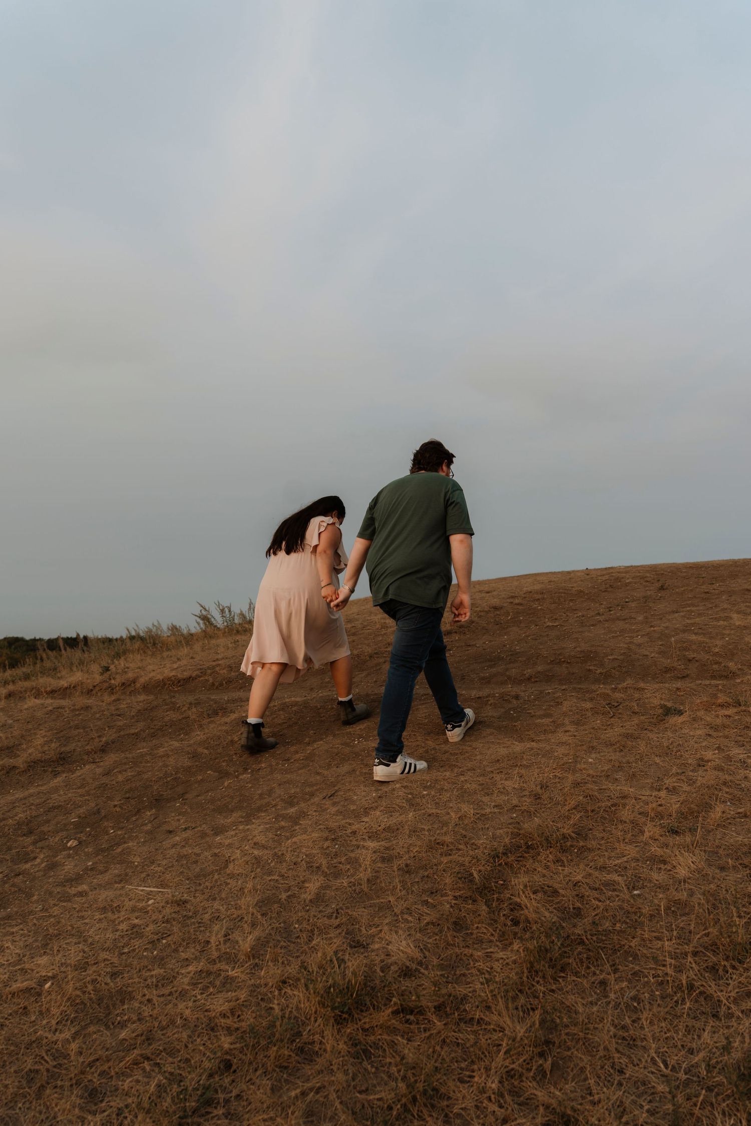 A couple holds hands while walking up a dry grassy hill under a cloudy sky.