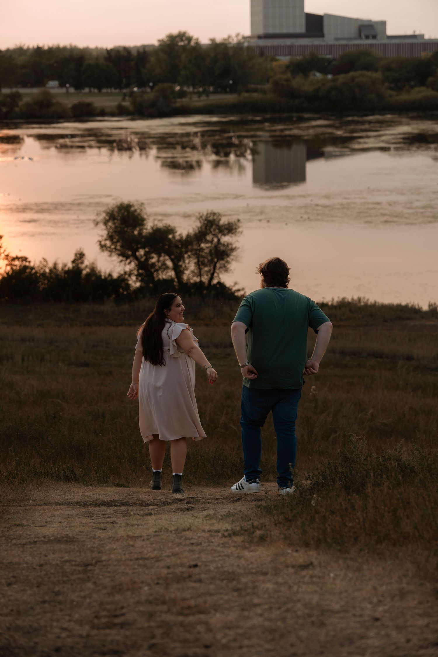 Couple walking hand in hand toward a serene river at sunset, surrounded by golden light and lush greenery.