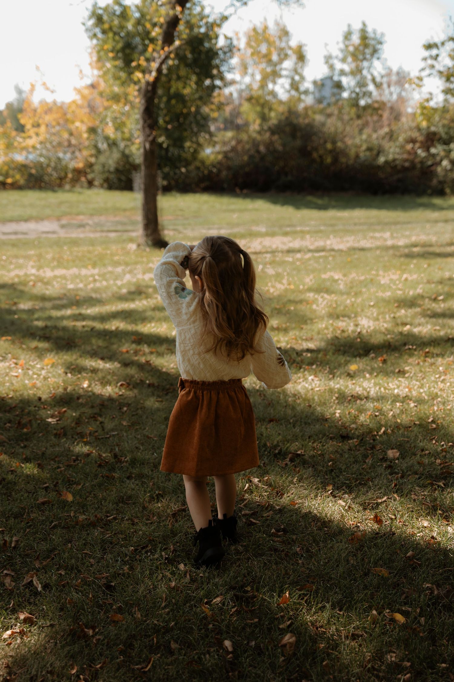 A young girl in a rust skirt and white sweater walks through a sunlit autumn park with fallen leaves.