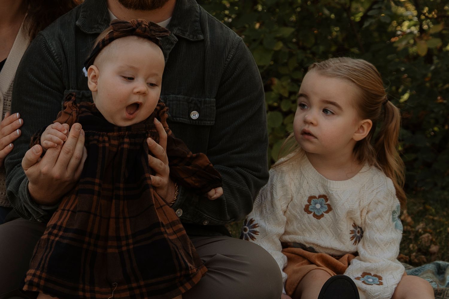 A baby in a plaid dress and a young girl in a floral sweater sit with an adult outdoors in autumn.
