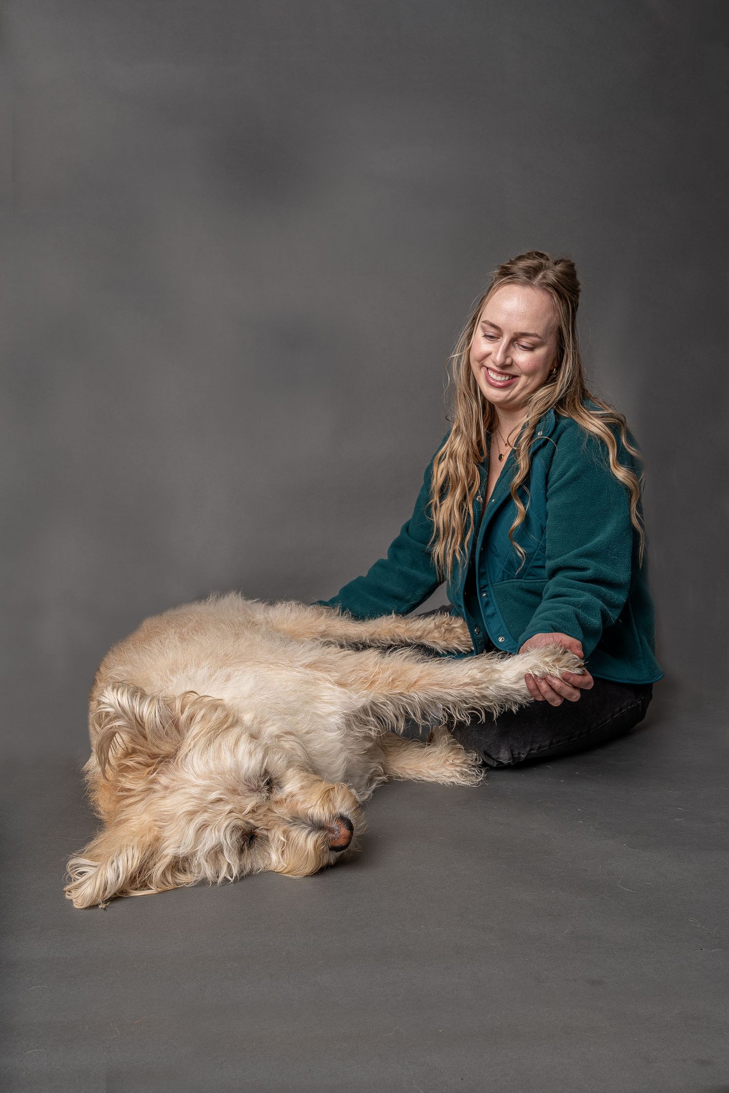 Smiling blonde woman in teal jacket sitting on floor with large fluffy cream-colored dog lying across her lap.