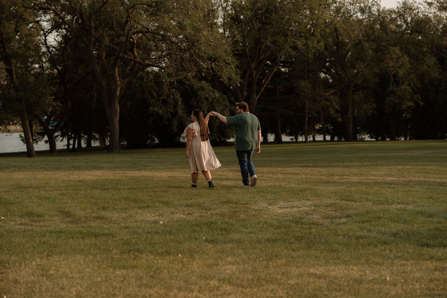 Couple holding hands dancing in a lush green park surrounded by tall trees on a cloudy day.
