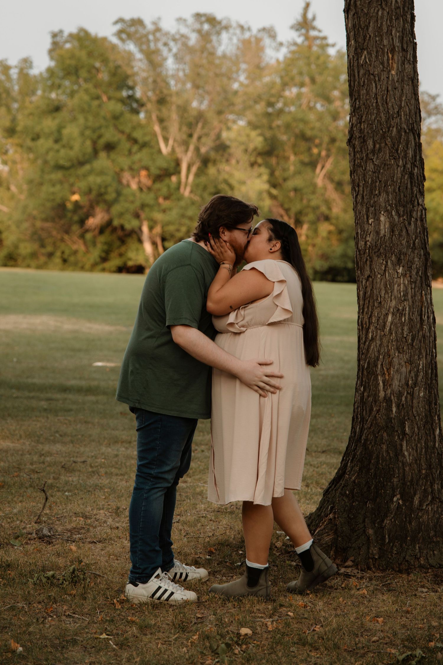 Couple sharing a romantic kiss near a tree in a park, woman in pink dress with visible baby bump.