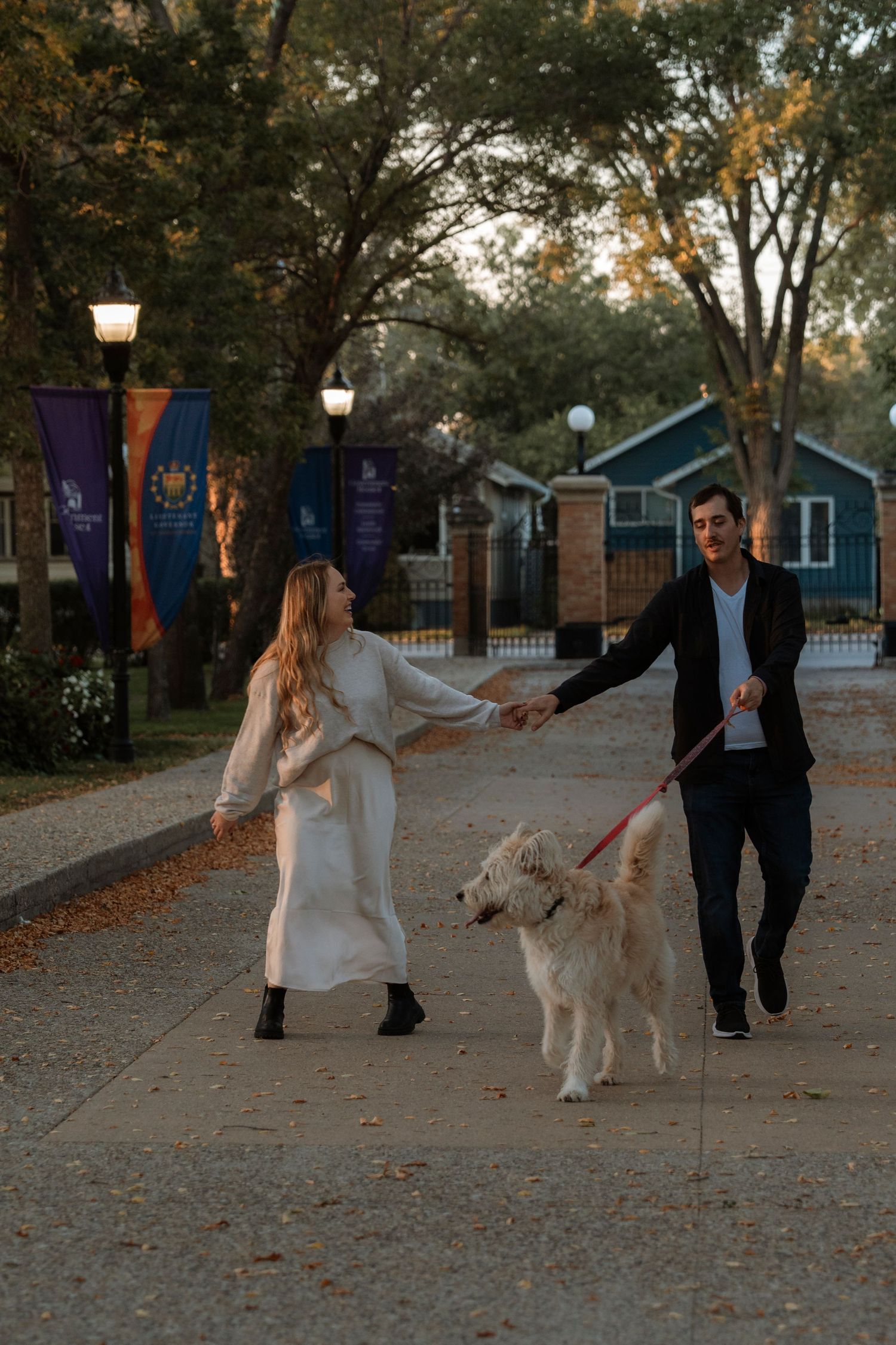 Couple walking a golden retriever on a tree-lined sidewalk at dusk, woman in white dress holding mans hand.
