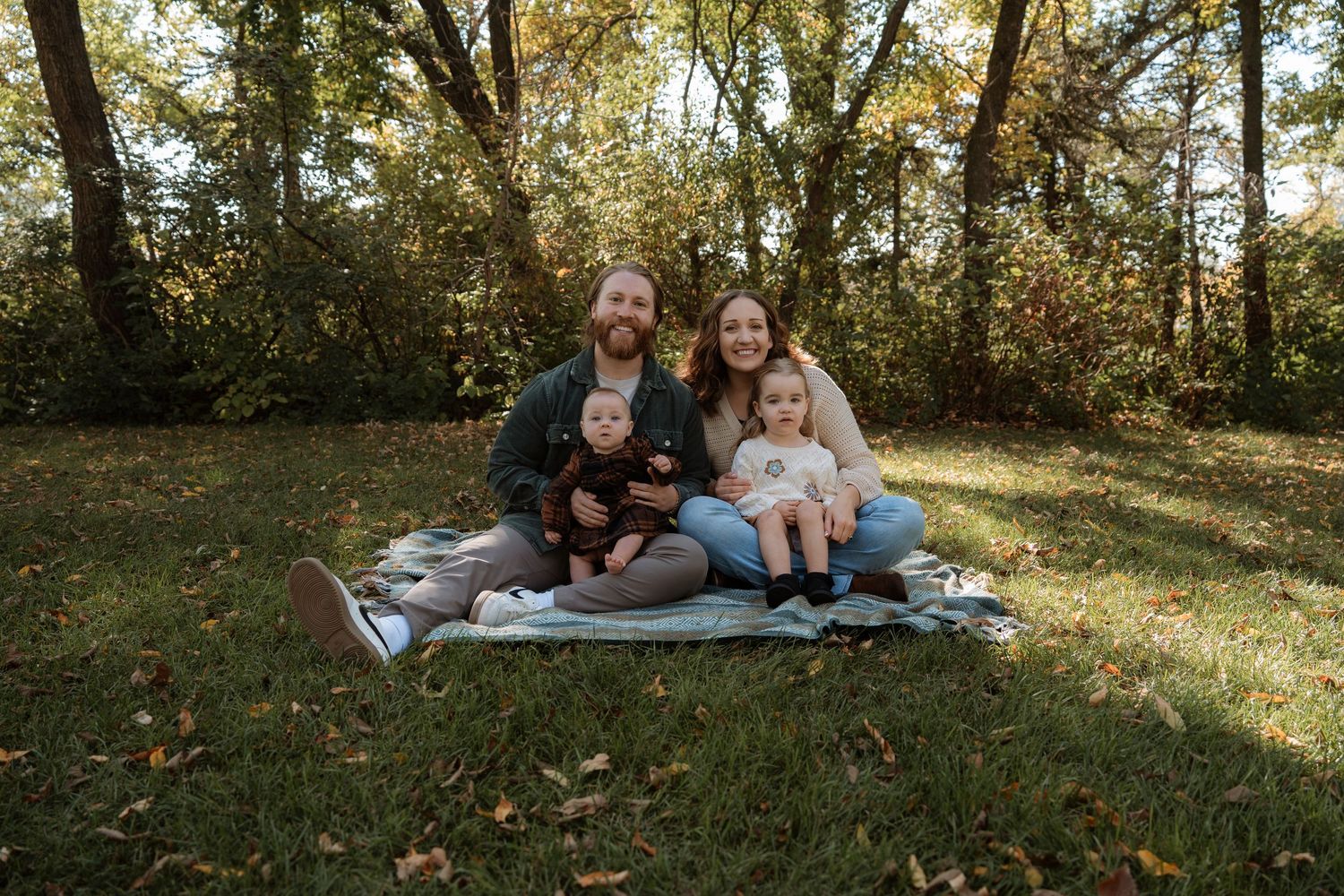 Family of four sitting on a blanket in a park during autumn, smiling together surrounded by colorful fall foliage.