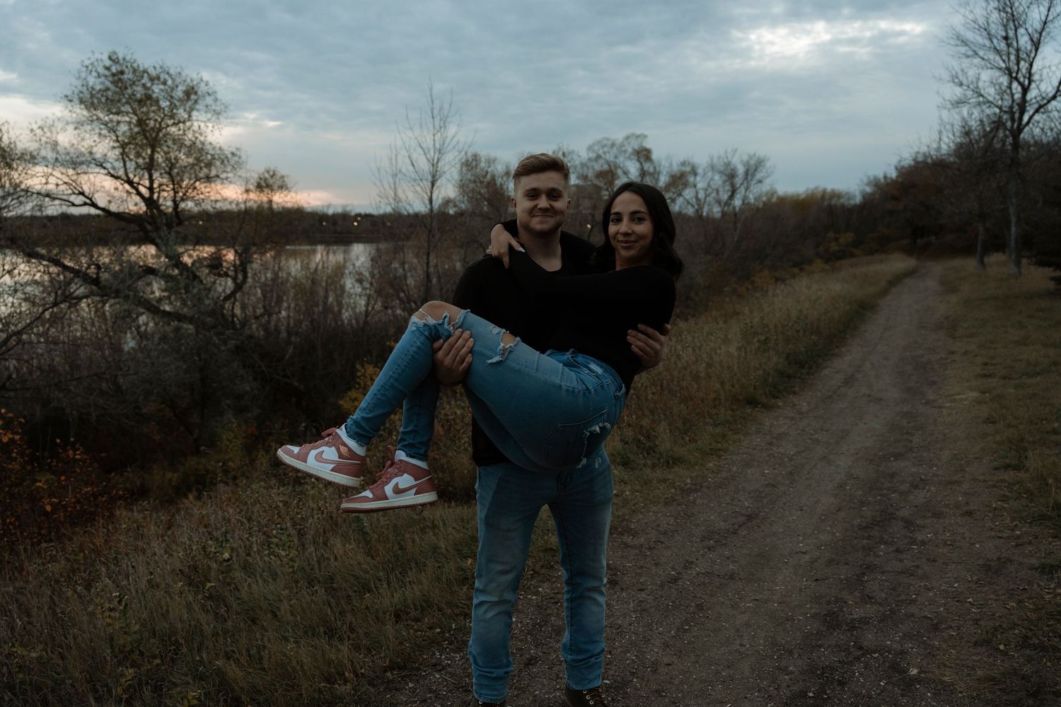 A happy young couple poses together on a rural dirt path at dusk near a river.