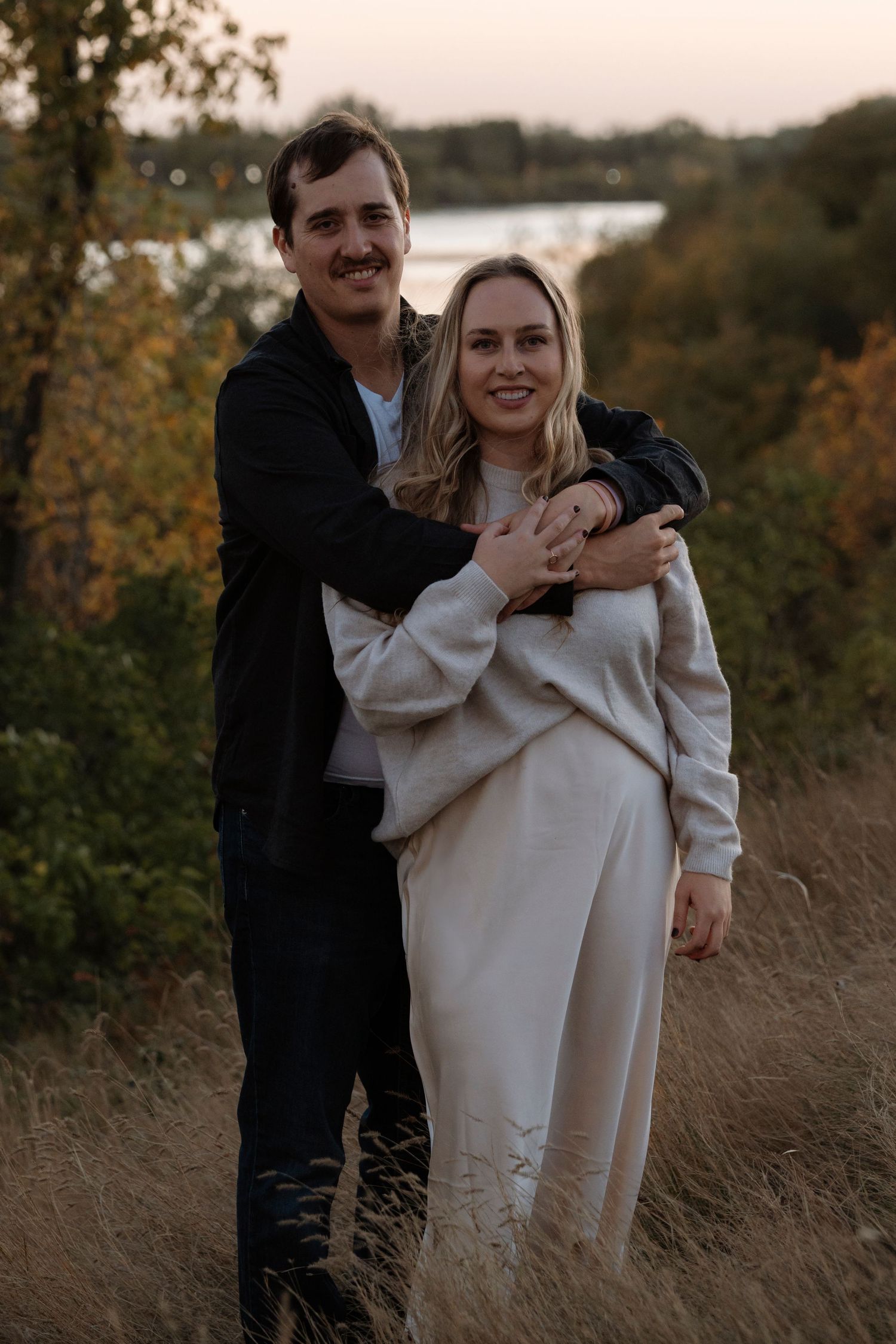 A smiling couple embraces outdoors in autumn, with a lake and colorful foliage in the background at dusk.