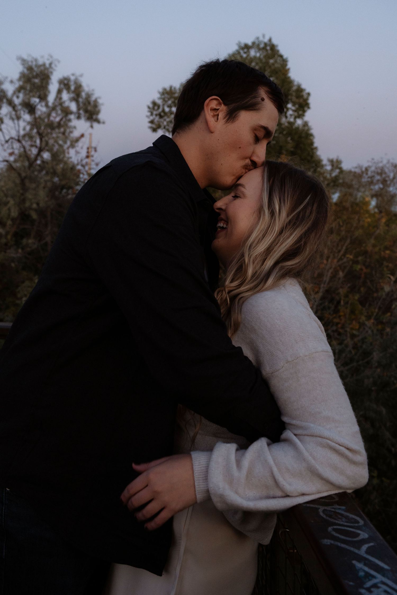 A couple shares a romantic kiss at dusk outdoors, the woman in a white sweater embraced by a man in a black jacket.