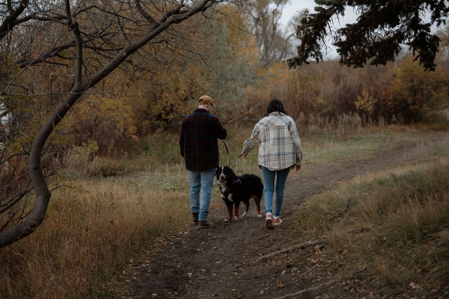 Two people walking a black dog on a dirt trail through autumn foliage in a park.