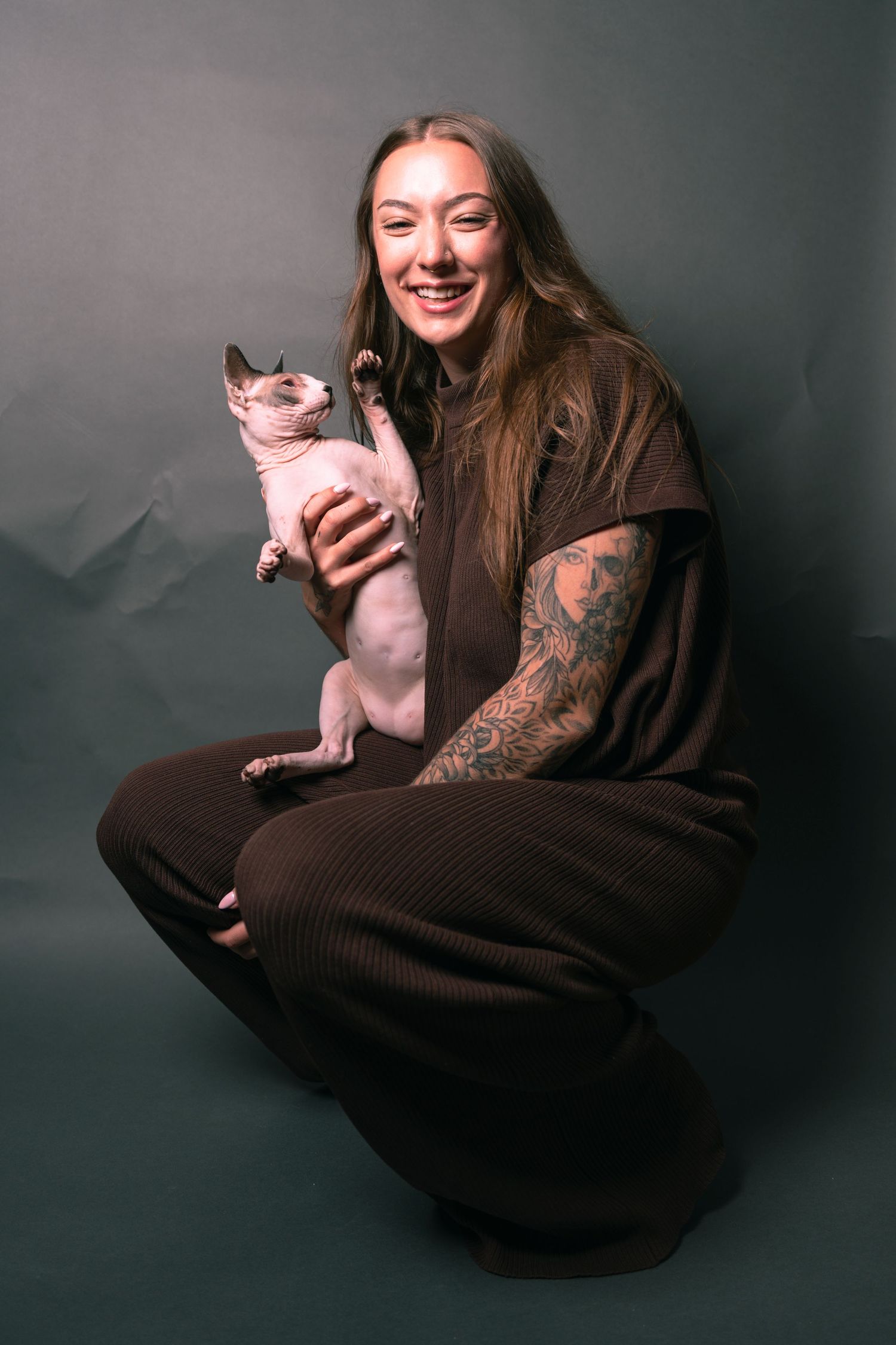 Smiling woman with tattoos crouching in brown outfit, holding a Sphynx cat against a dark gray background.