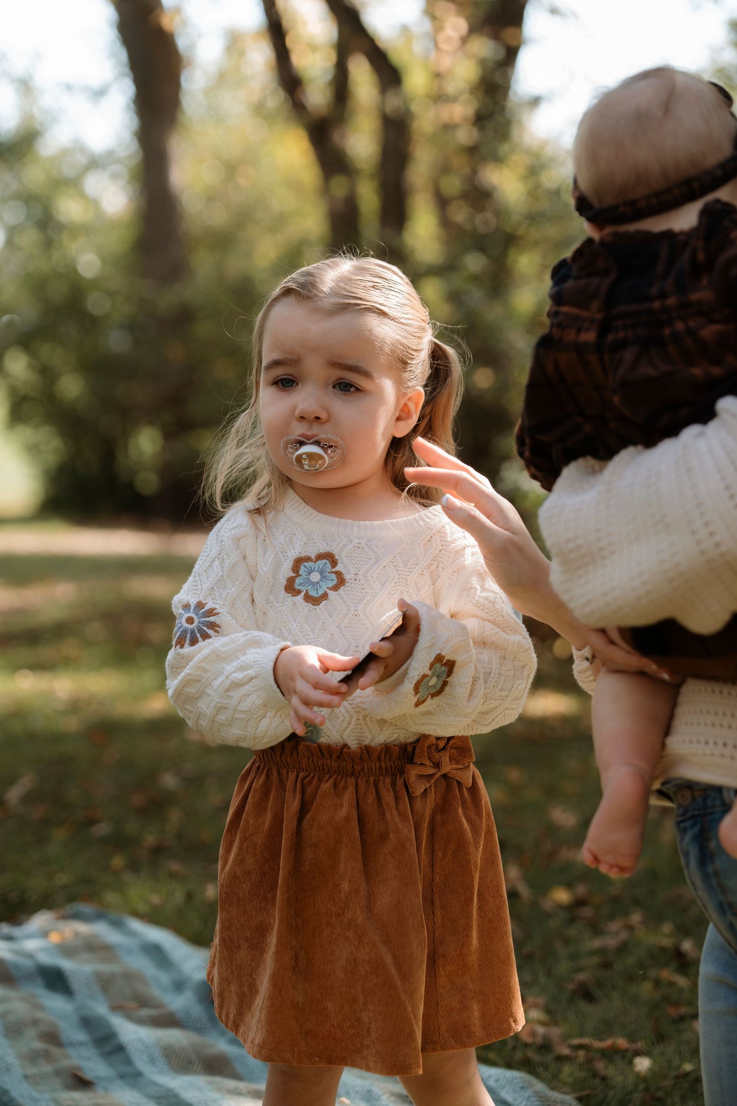 Toddler girl in floral sweater and brown skirt with pacifier stands outdoors on blanket in autumn park.