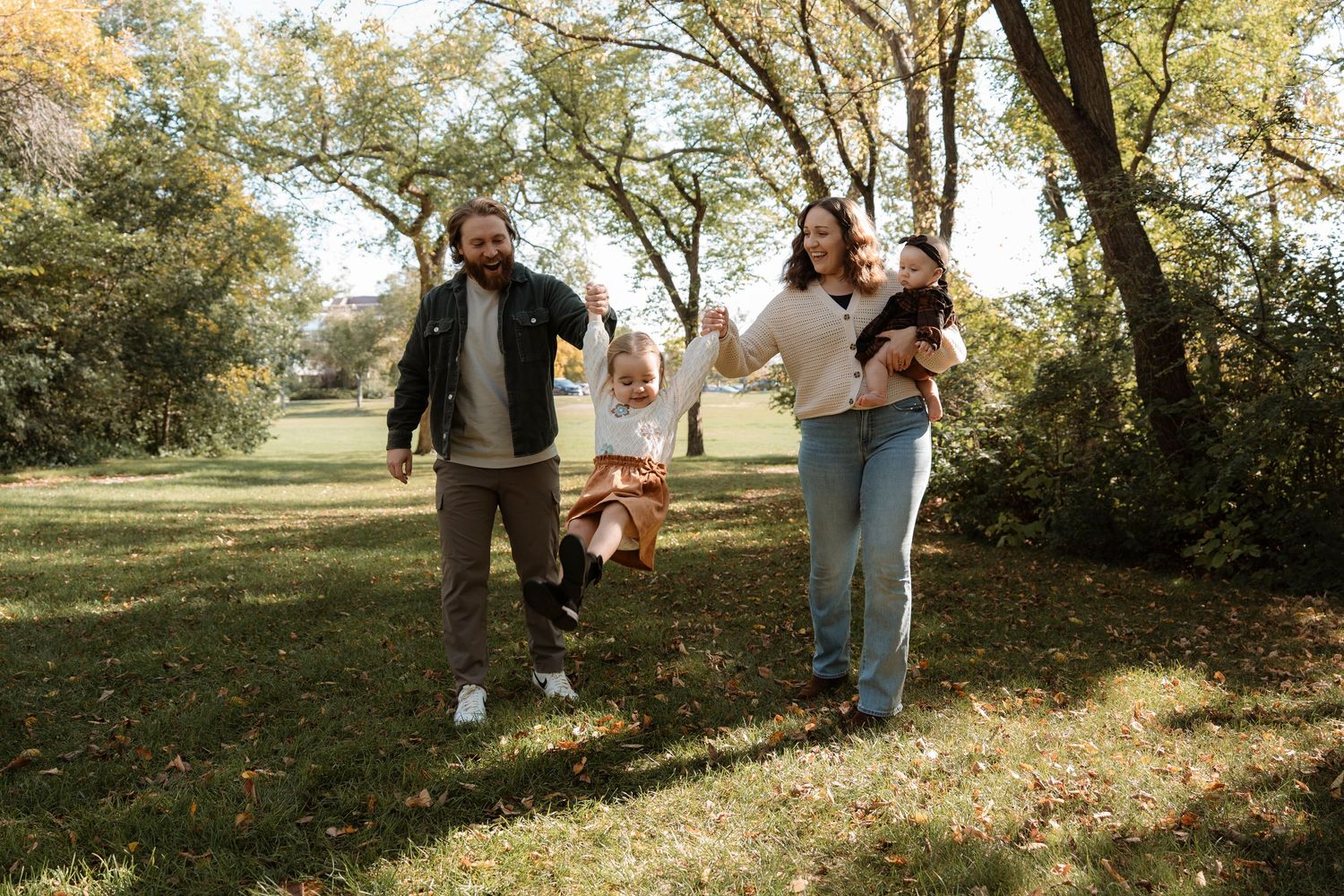 Family with two children playing outdoors in a park during autumn, swinging toddler between parents.