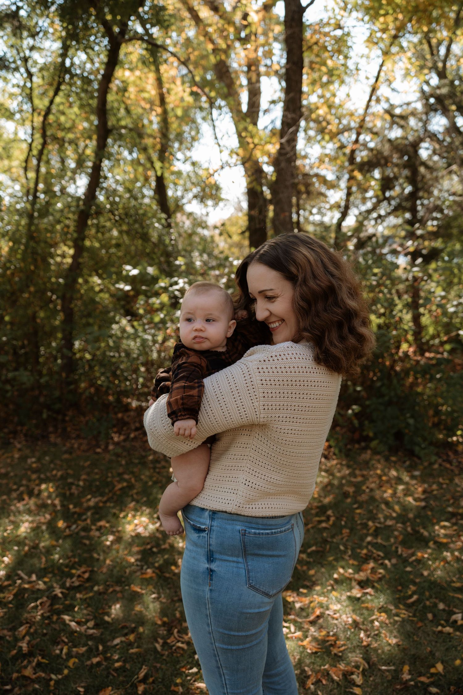 A smiling mother holds her baby in a sunlit autumn forest with fallen leaves and golden trees.