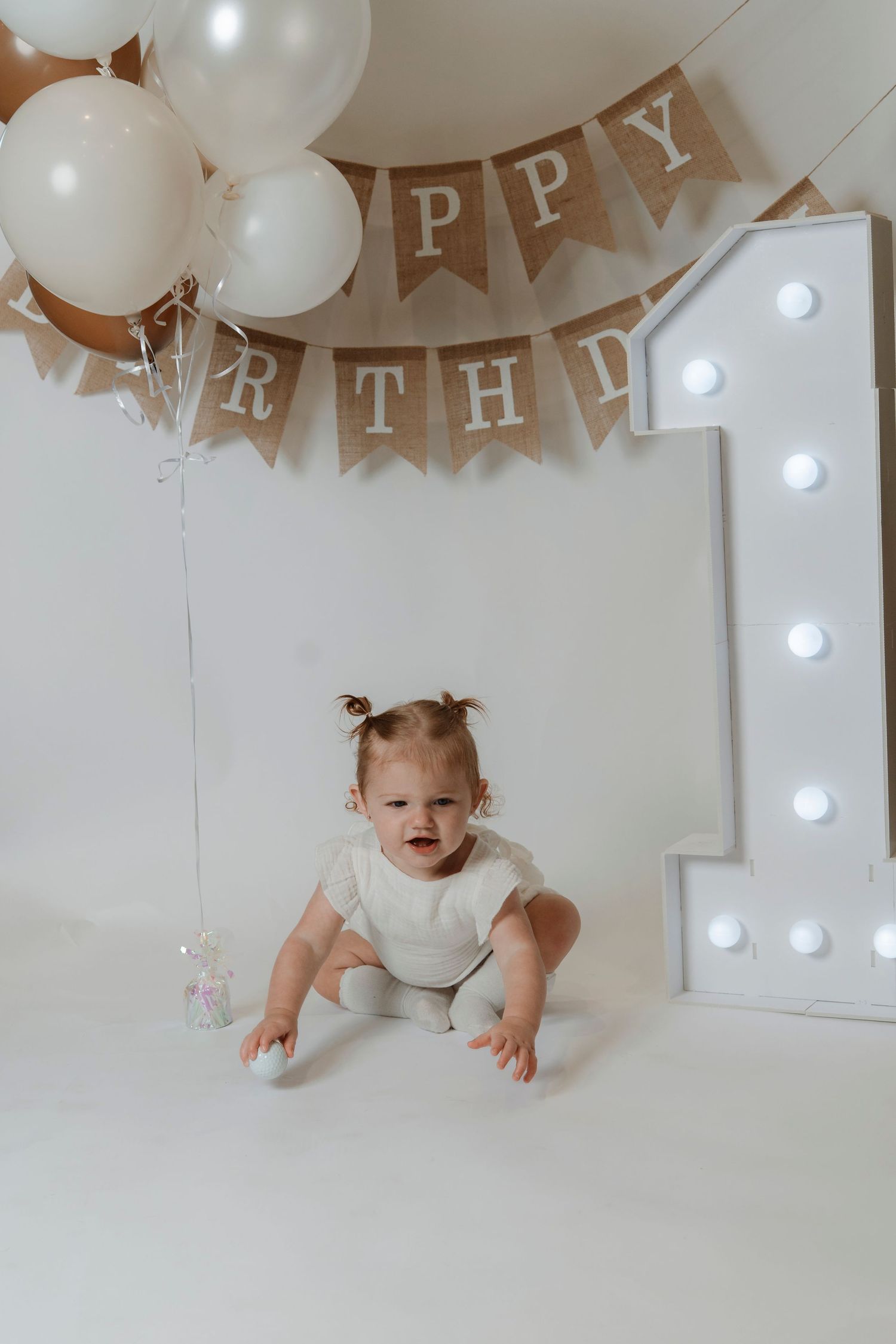 Baby girl in white onesie sits on floor at first birthday party with balloons and Happy Birthday banner.