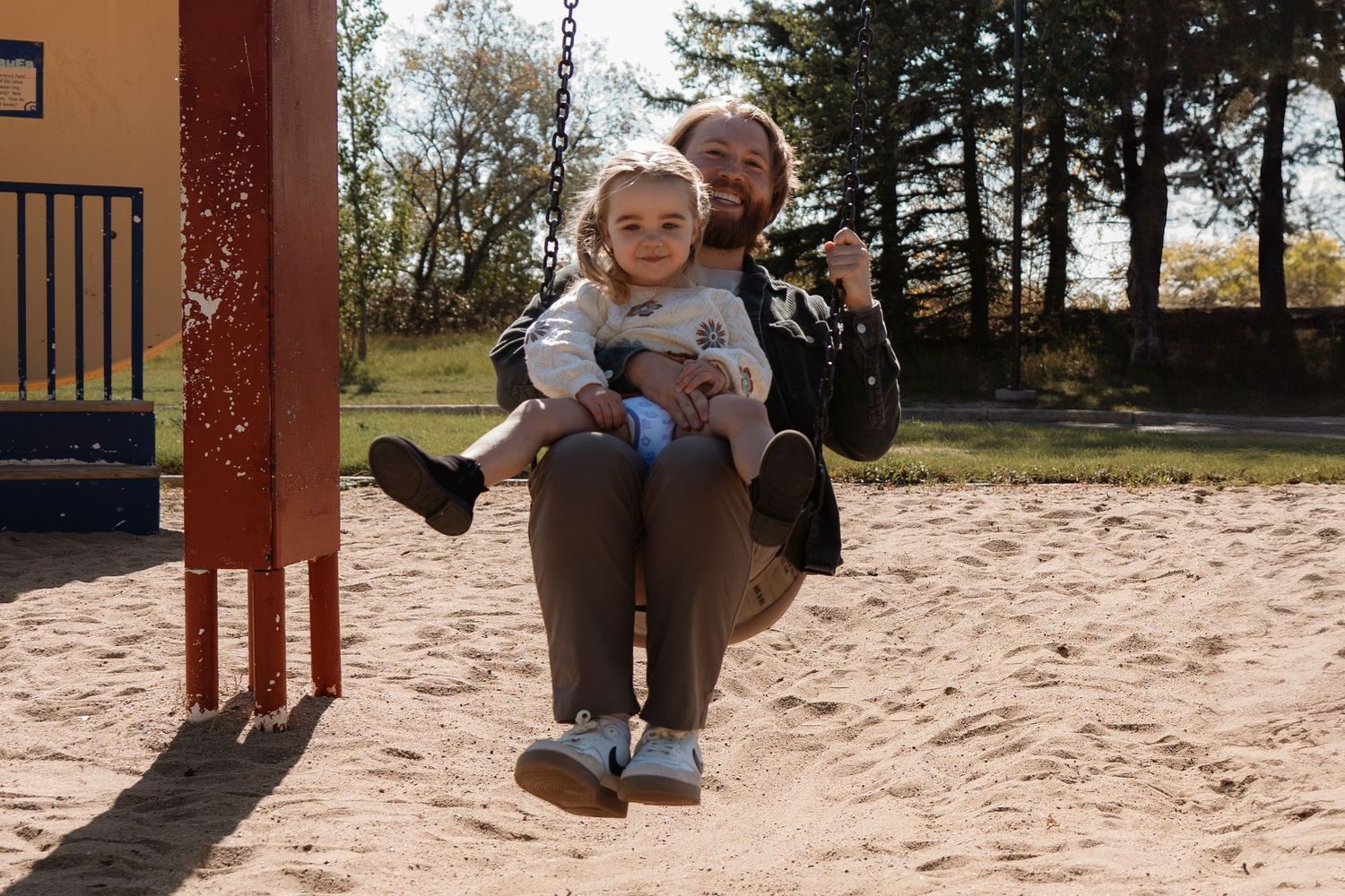 Adult and toddler sharing a swing at a sandy playground, surrounded by trees on a sunny day.