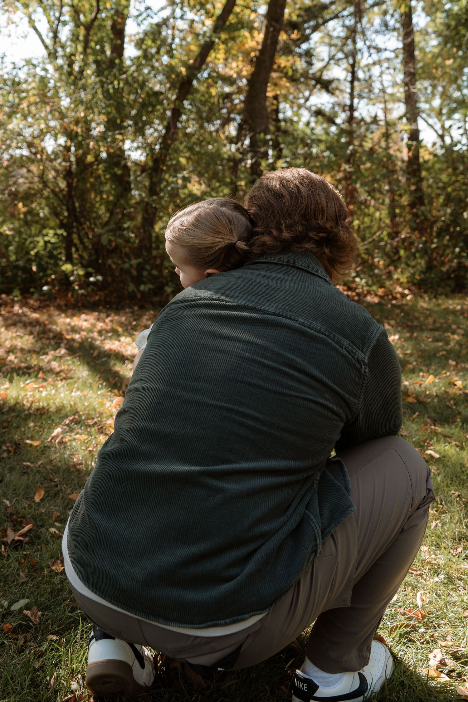 A parent crouches to embrace a child from behind in an autumn park, both facing away toward colorful trees.