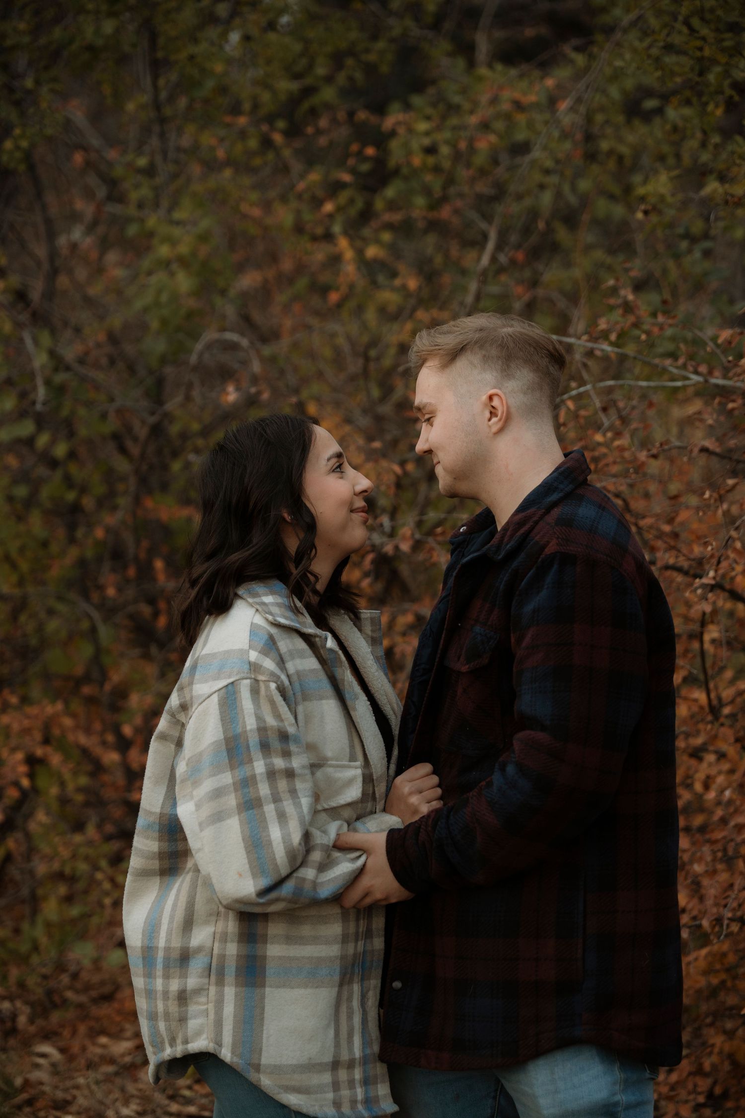 Couple holding hands and gazing at each other in autumn forest, wearing plaid shirts, surrounded by fall foliage.