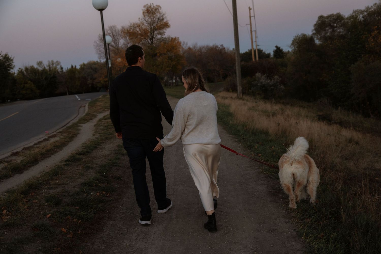 Couple walking hand in hand with a white dog on a leash along a dirt path at dusk.