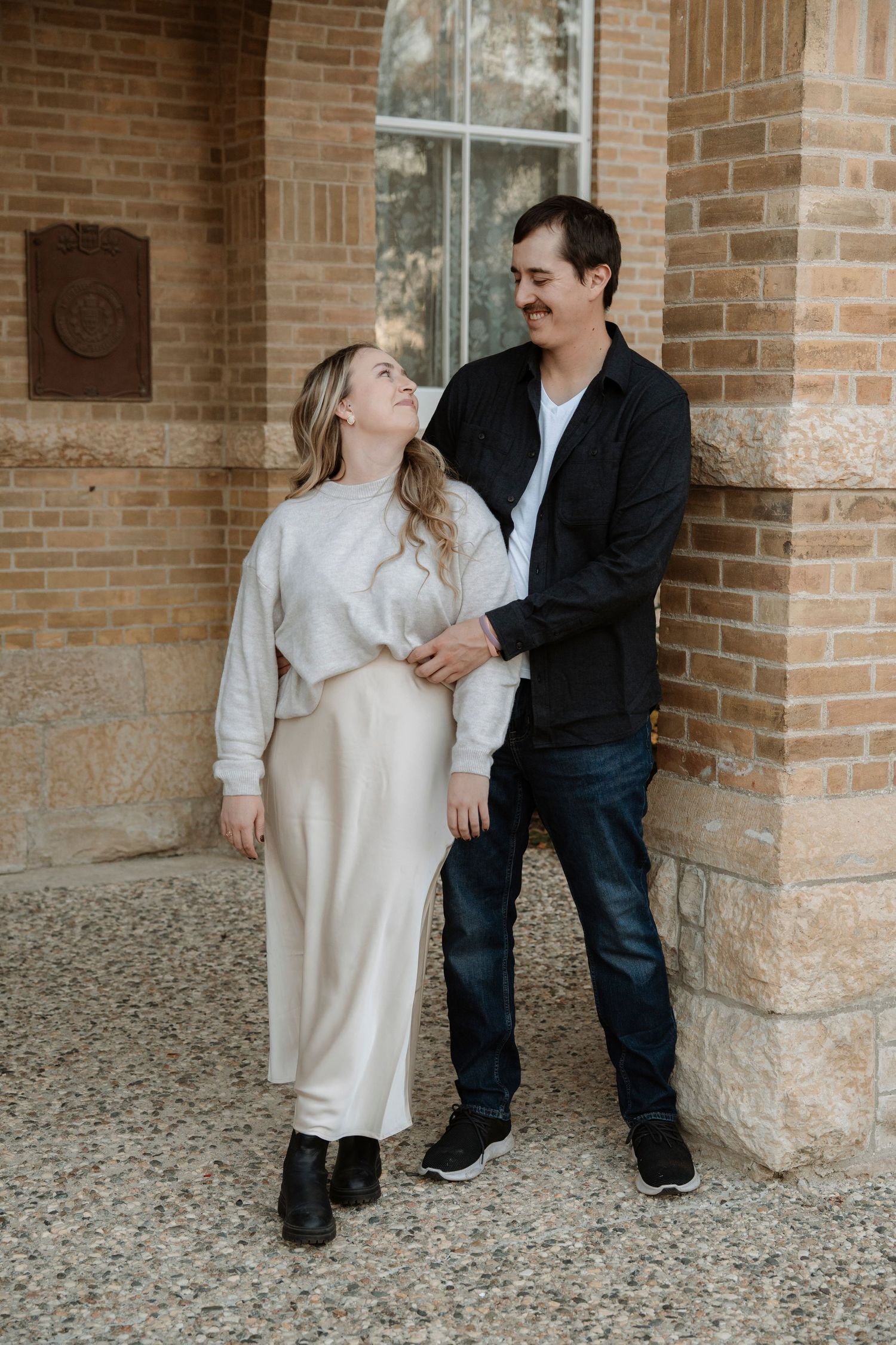 A couple shares a romantic moment leaning against a brick building, woman in cream skirt and man in black jacket.