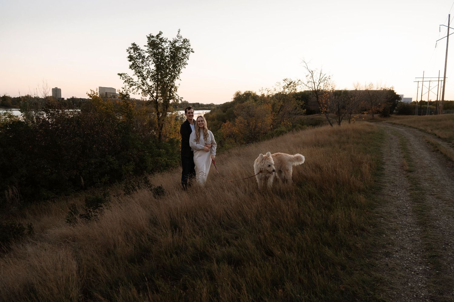 Couple in wedding attire walking with a white dog along a rural path at dusk with trees and power lines.