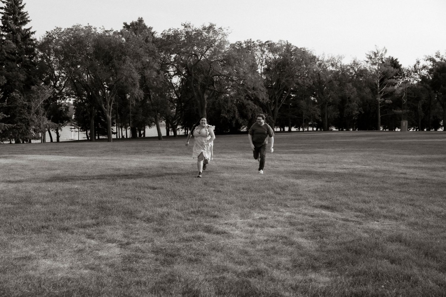 Two people running across a vast green park lawn with trees in the background, captured in black and white.