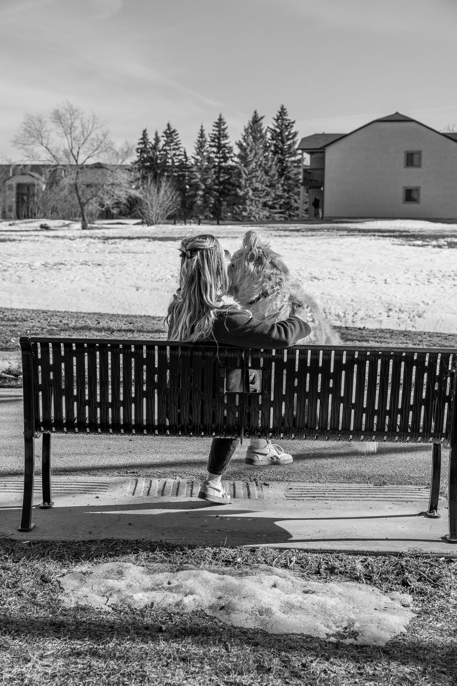 Two girls sit closely together on a park bench in winter, sharing a moment amid a snowy landscape with evergreen trees.