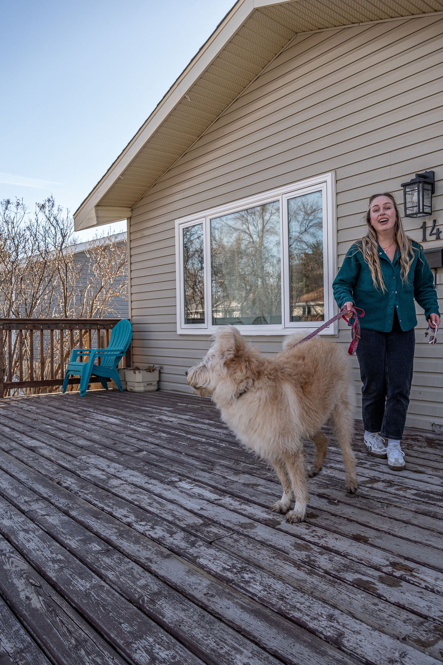 Woman in green shirt walking a fluffy golden dog on a leash across a wooden deck outside a house.