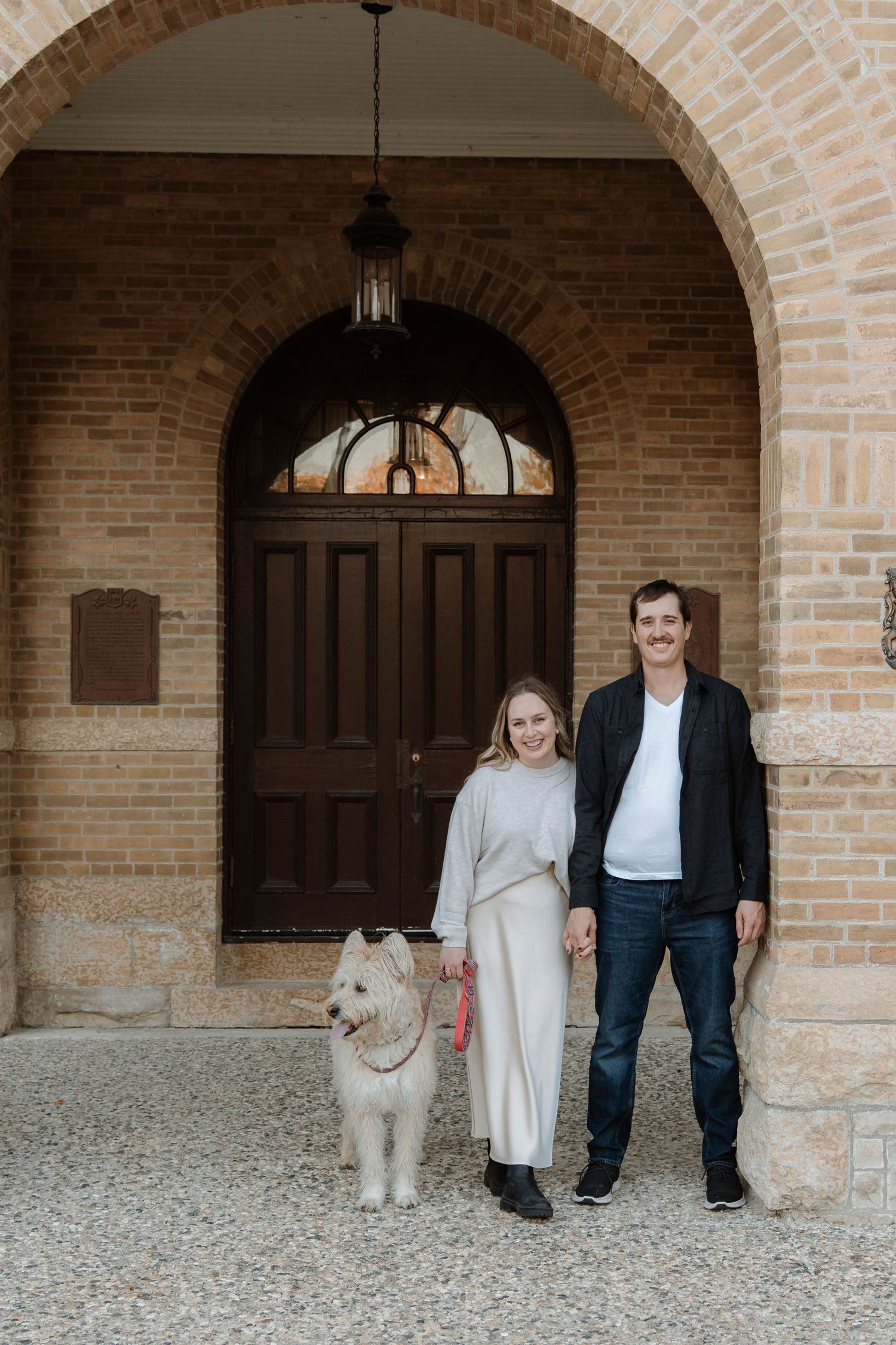 Couple holding hands with white dog standing before ornate brick archway and wooden double doors.