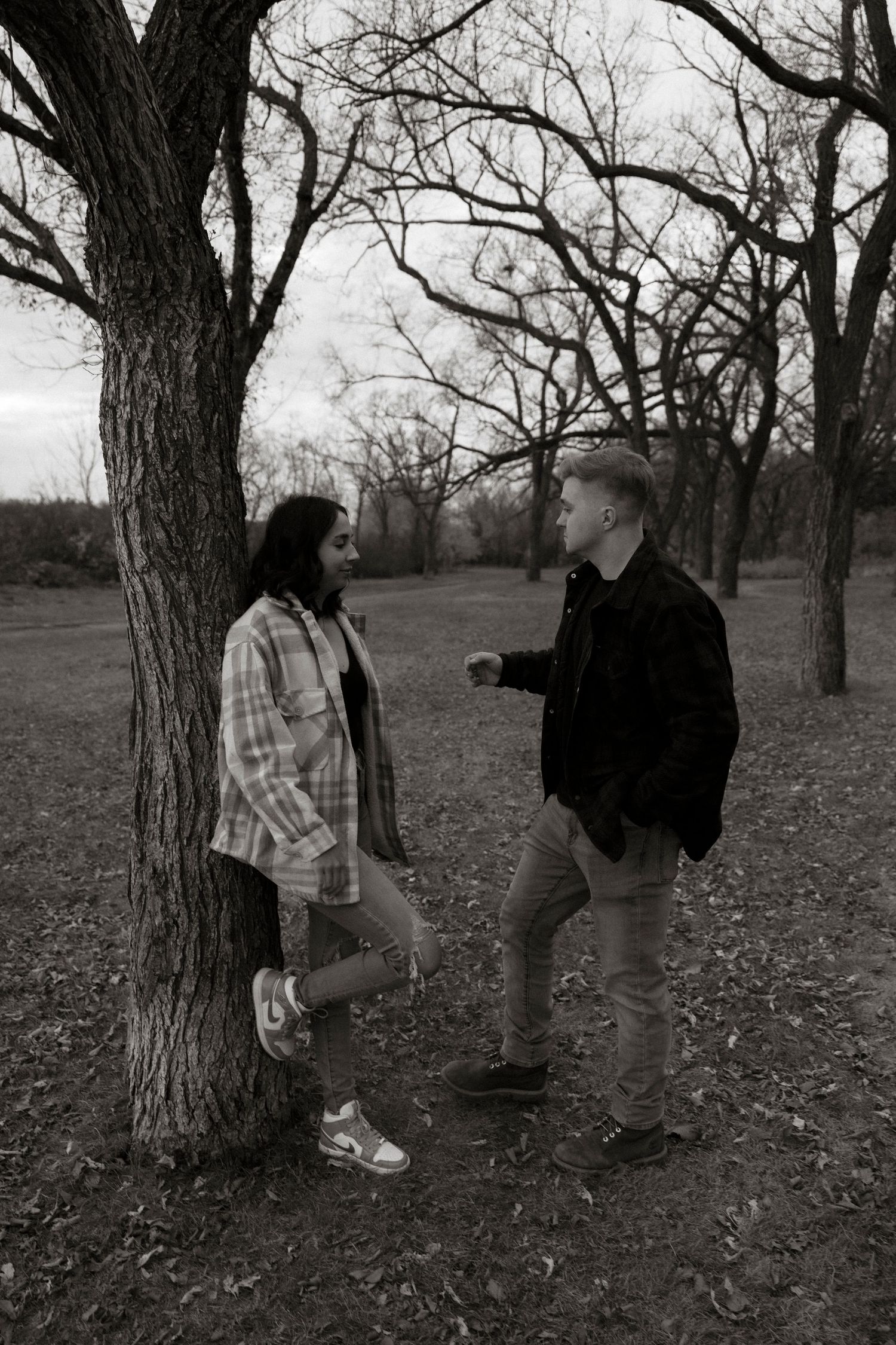Black and white photo of a young couple talking near a tree in a park during autumn.