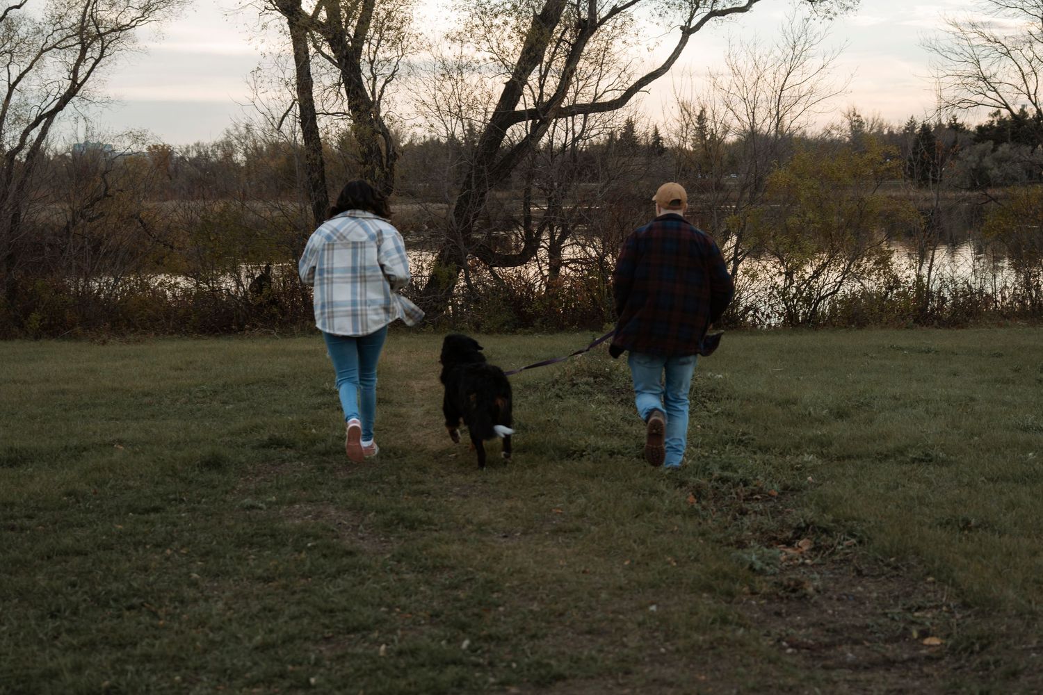Two people walking a black dog on a leash through a grassy park near a river on a cloudy autumn day.